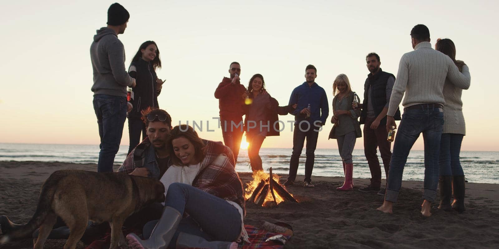 Happy Carefree Young Friends Having Fun And Drinking Beer By Bonefire On The Beach As The Sun Begins To Set