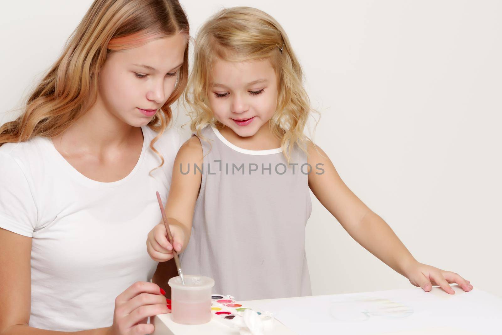 The older sister paints with the younger sister paints at the table on a piece of paper.