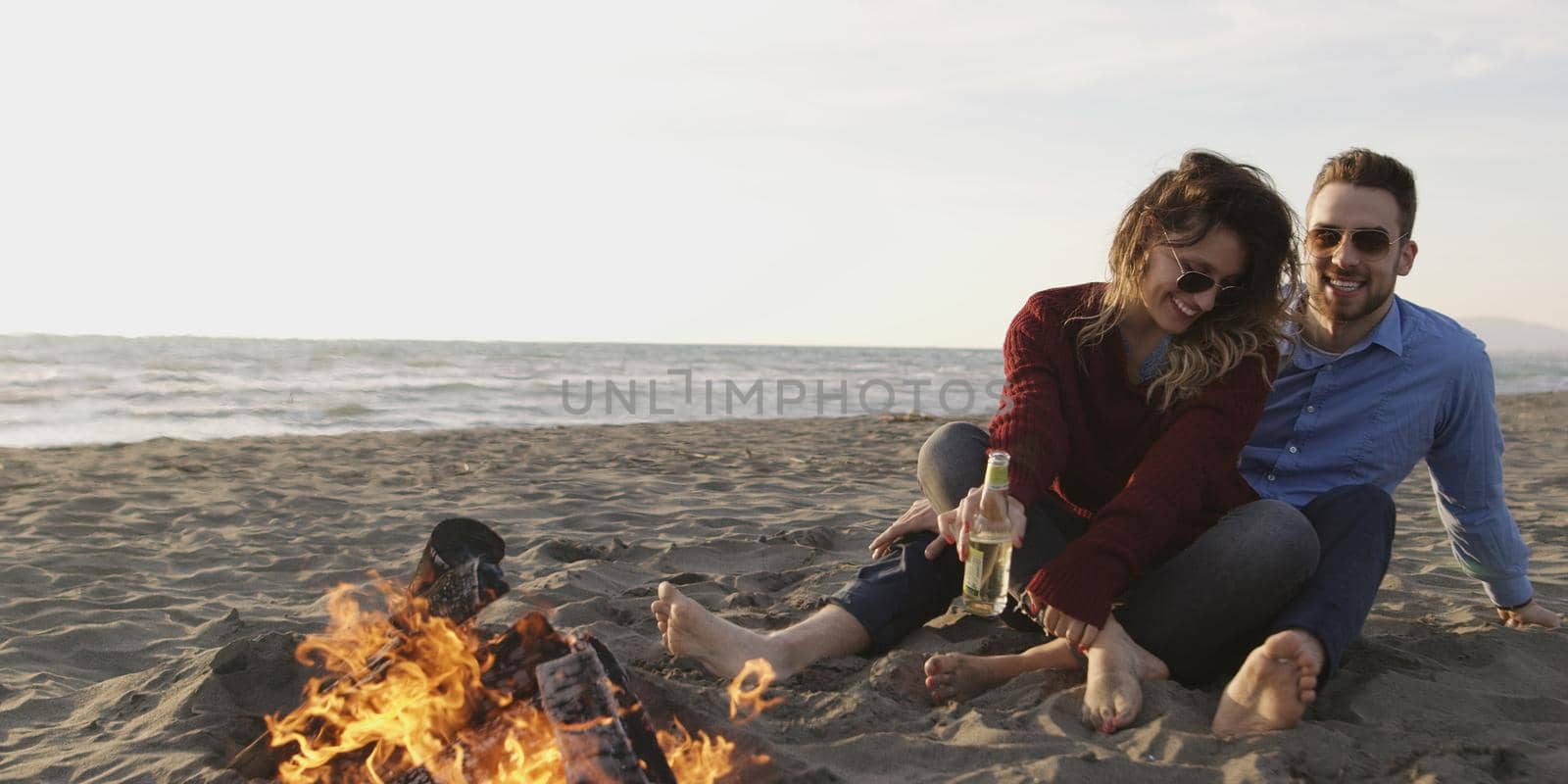 Young Couple Relaxing By The Fire, Drinking A Beer Or A Drink From The Bottle.