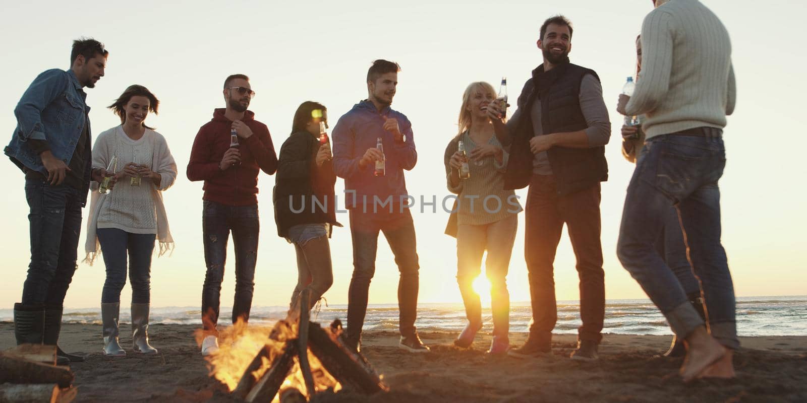 Happy Carefree Young Friends Having Fun And Drinking Beer By Bonefire On The Beach As The Sun Begins To Set