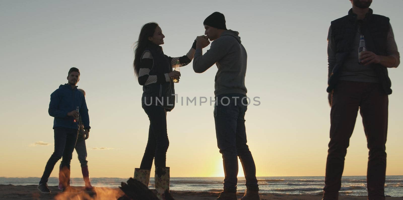 Happy Carefree Young Friends Having Fun And Drinking Beer By Bonefire On The Beach As The Sun Begins To Set