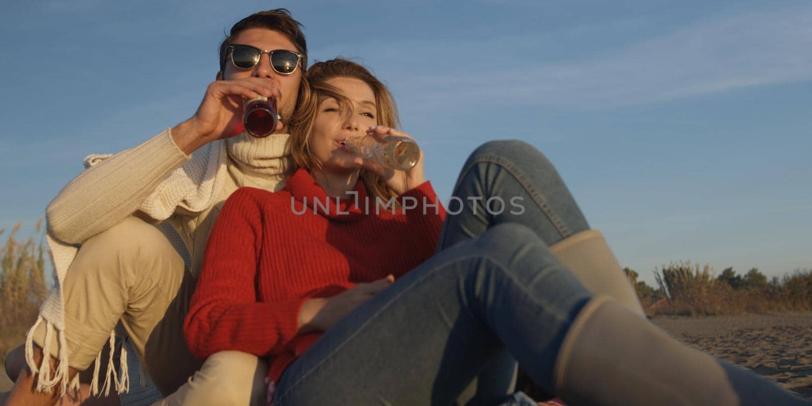 Young Couple Relaxing By The Fire, Drinking A Beer Or A Drink From The Bottle.