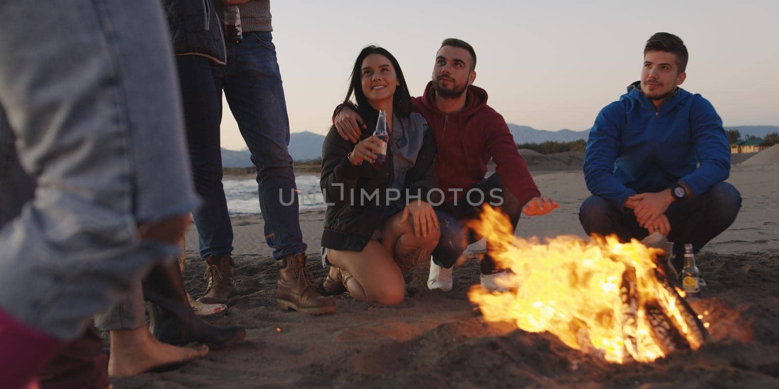 Happy Carefree Young Friends Having Fun And Drinking Beer By Bonefire On The Beach As The Sun Begins To Set