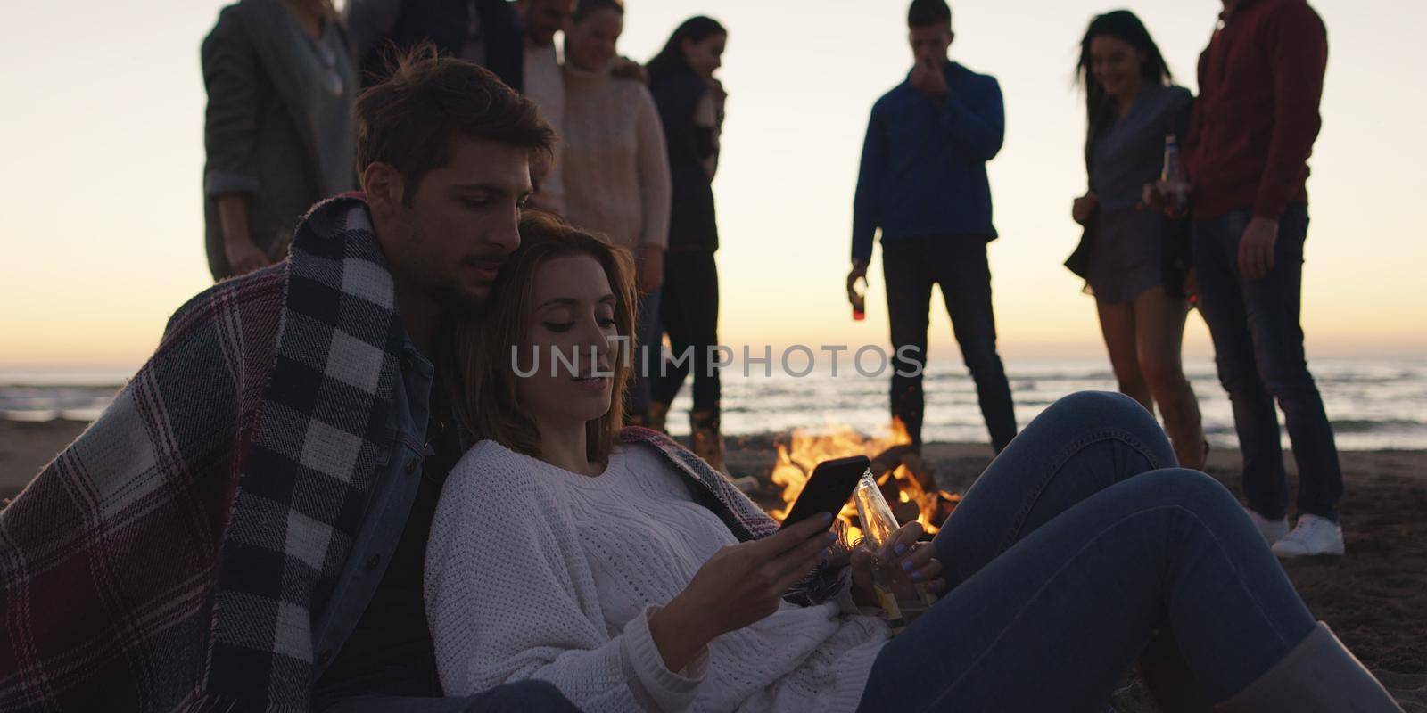 Couple using cell phone during beach party with friends drinking beer and having fun