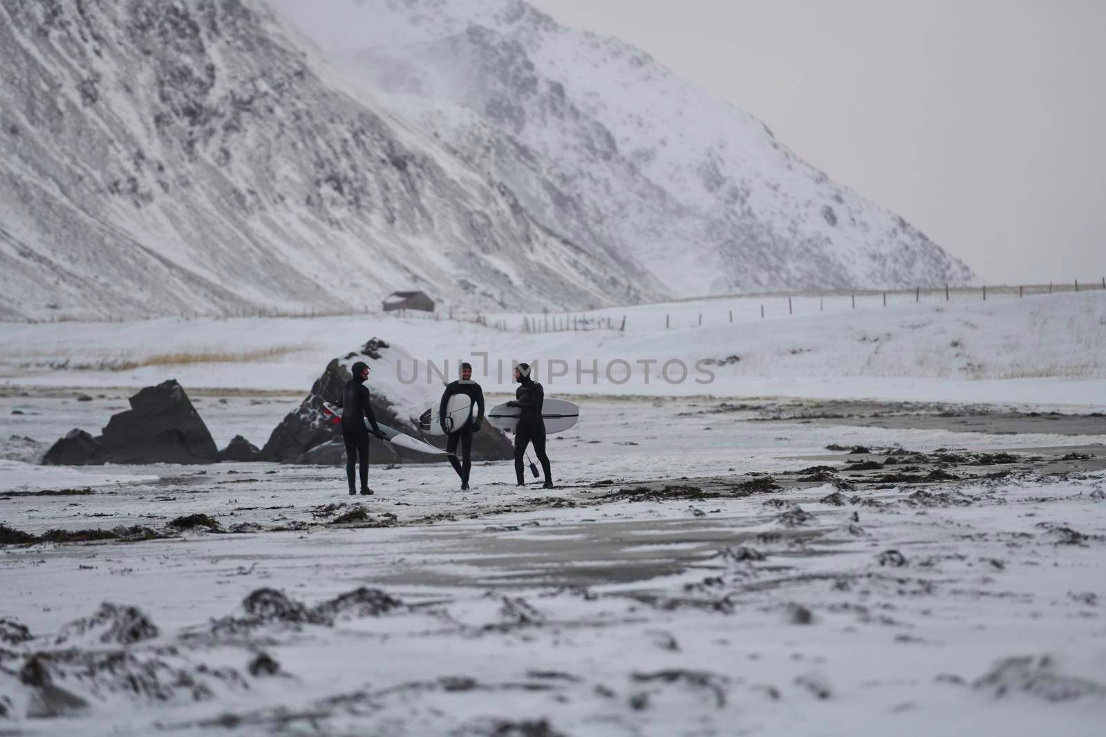 Authentic local Arctic surfers going by beach after surfing in Norwegian sea at snow capped mountain background in winter. Unstad, Norwegian village on Lofoten islands. Norwegian sea coastline. Winter water activities extreme sport