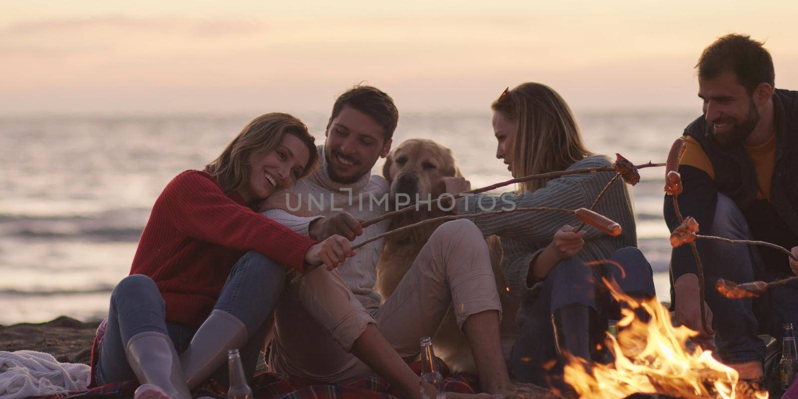 Group of young friends sitting by the fire late at night, grilling sausages and drinking beer, talking and having fun