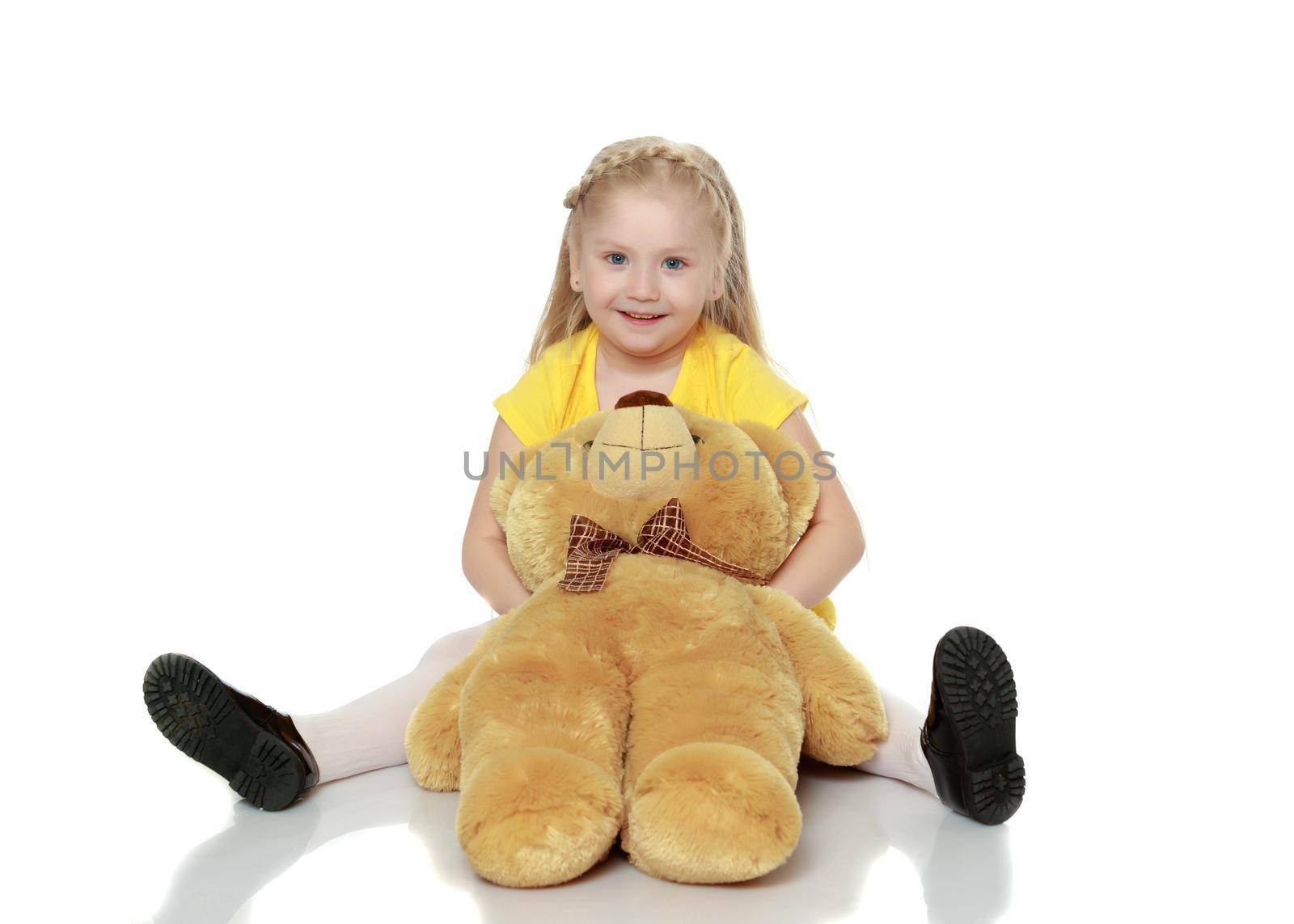 A lovely little round-faced blonde girl, with very long beautiful hair, in short skirts and yellow jerseys.She hugs a big teddy bear.Isolated on white background.