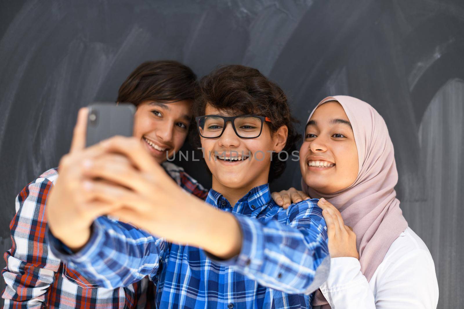 Group of arab teens taking selfie photo on smart phone with black chalkboard in background. Selective focus  by dotshock