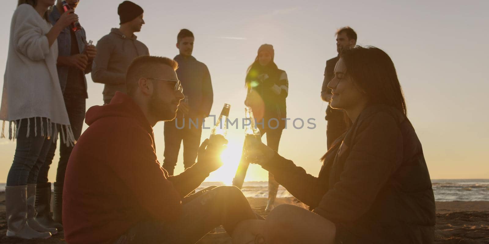 Happy Carefree Young Friends Having Fun And Drinking Beer By Bonefire On The Beach As The Sun Begins To Set