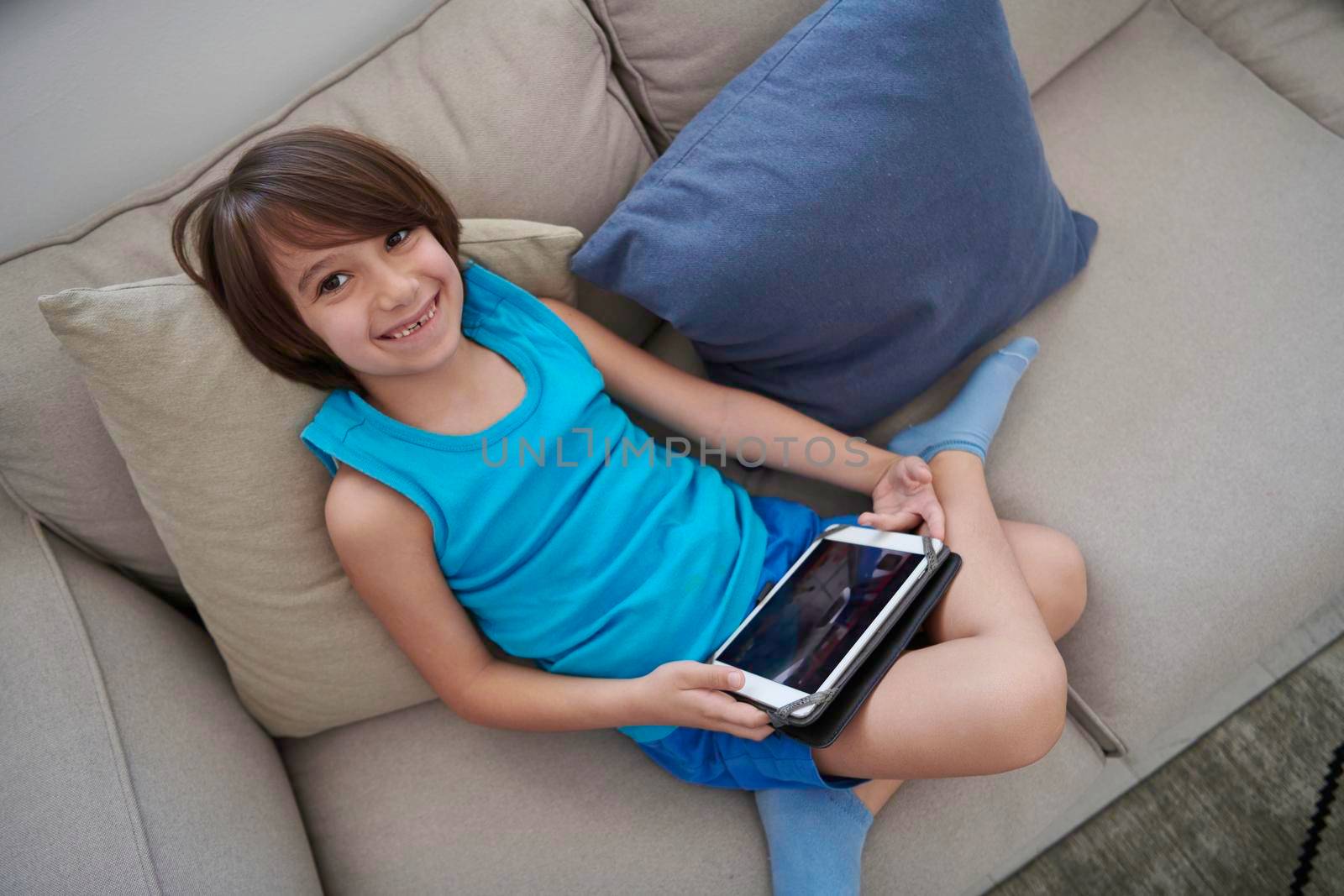 Little Arabian boy sitting on sofa and playing game on digital tablet. Portrait of a young arab child at home watching cartoon on the laptop. Modern kid and online education elearning technology.