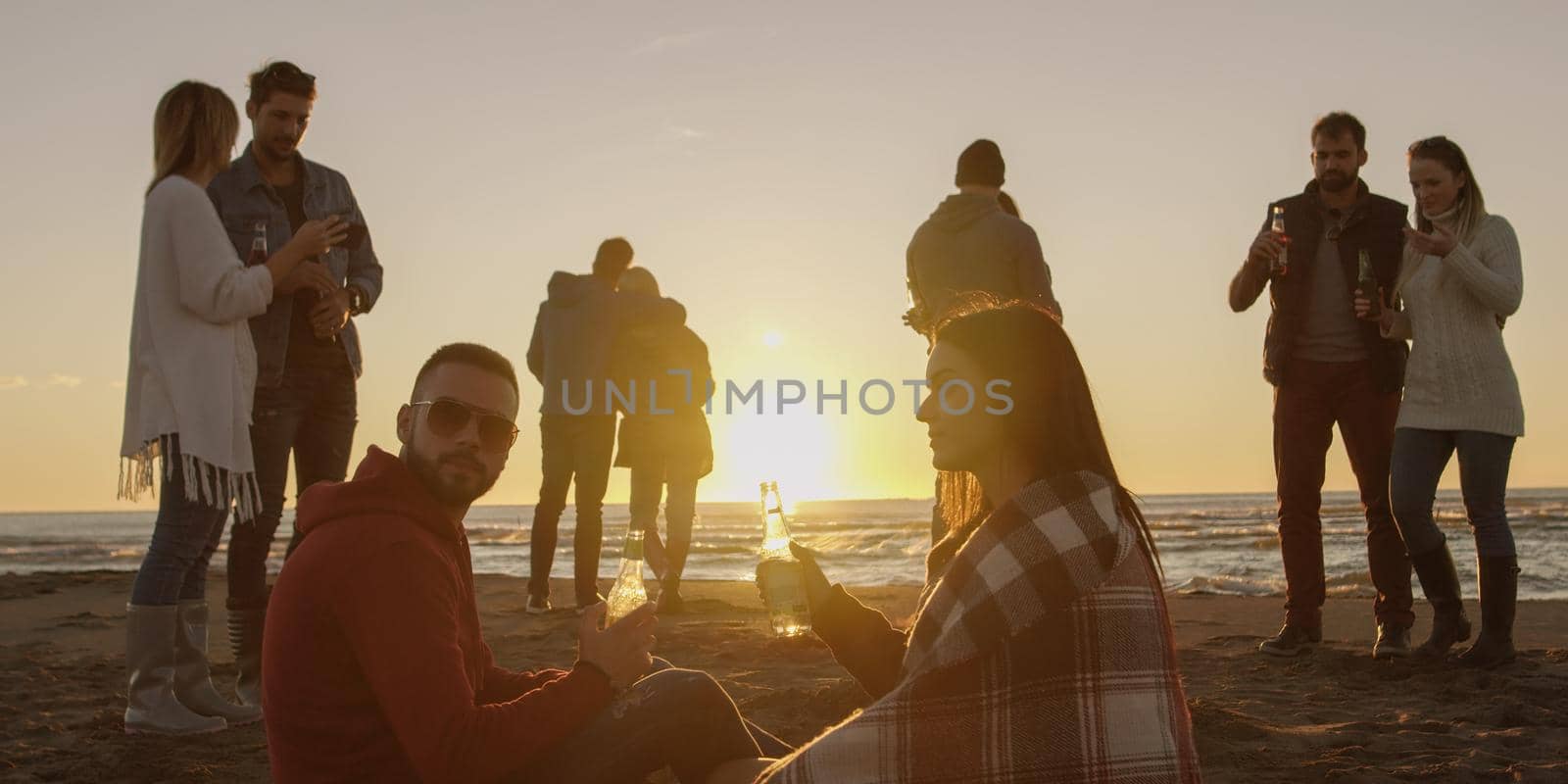 Happy Carefree Young Friends Having Fun And Drinking Beer By Bonefire On The Beach As The Sun Begins To Set