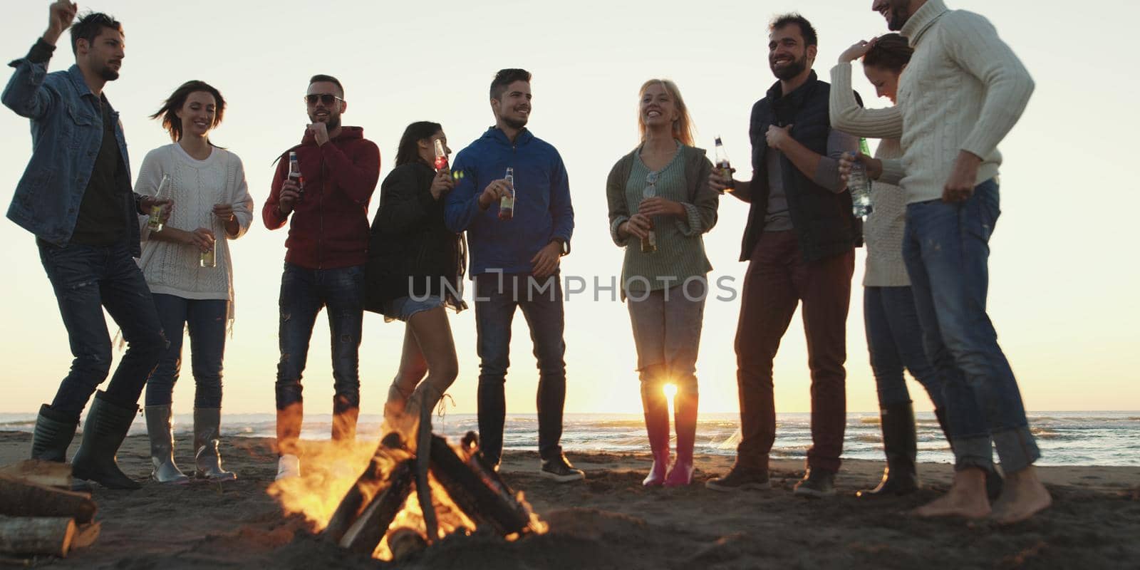 Happy Carefree Young Friends Having Fun And Drinking Beer By Bonefire On The Beach As The Sun Begins To Set