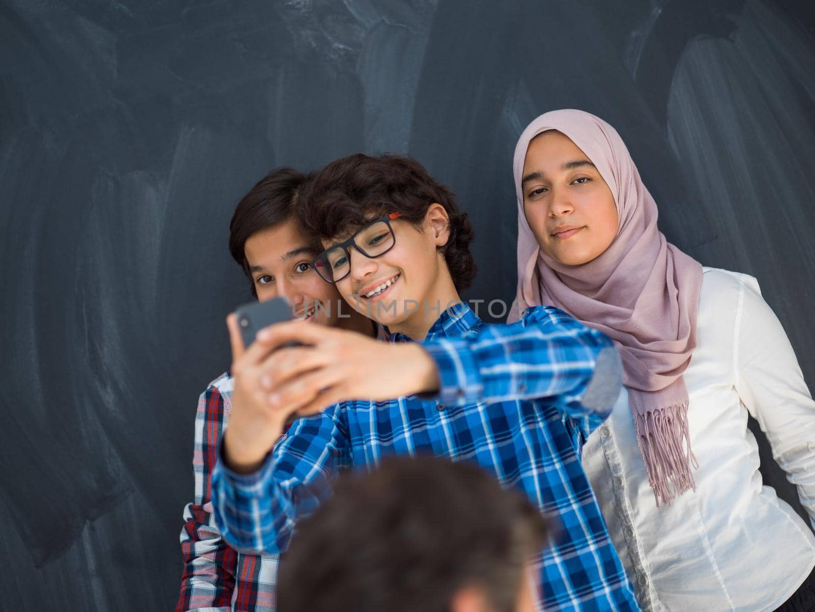 group of arab teens taking selfie photo on smart phone with black chalkboard in background by dotshock