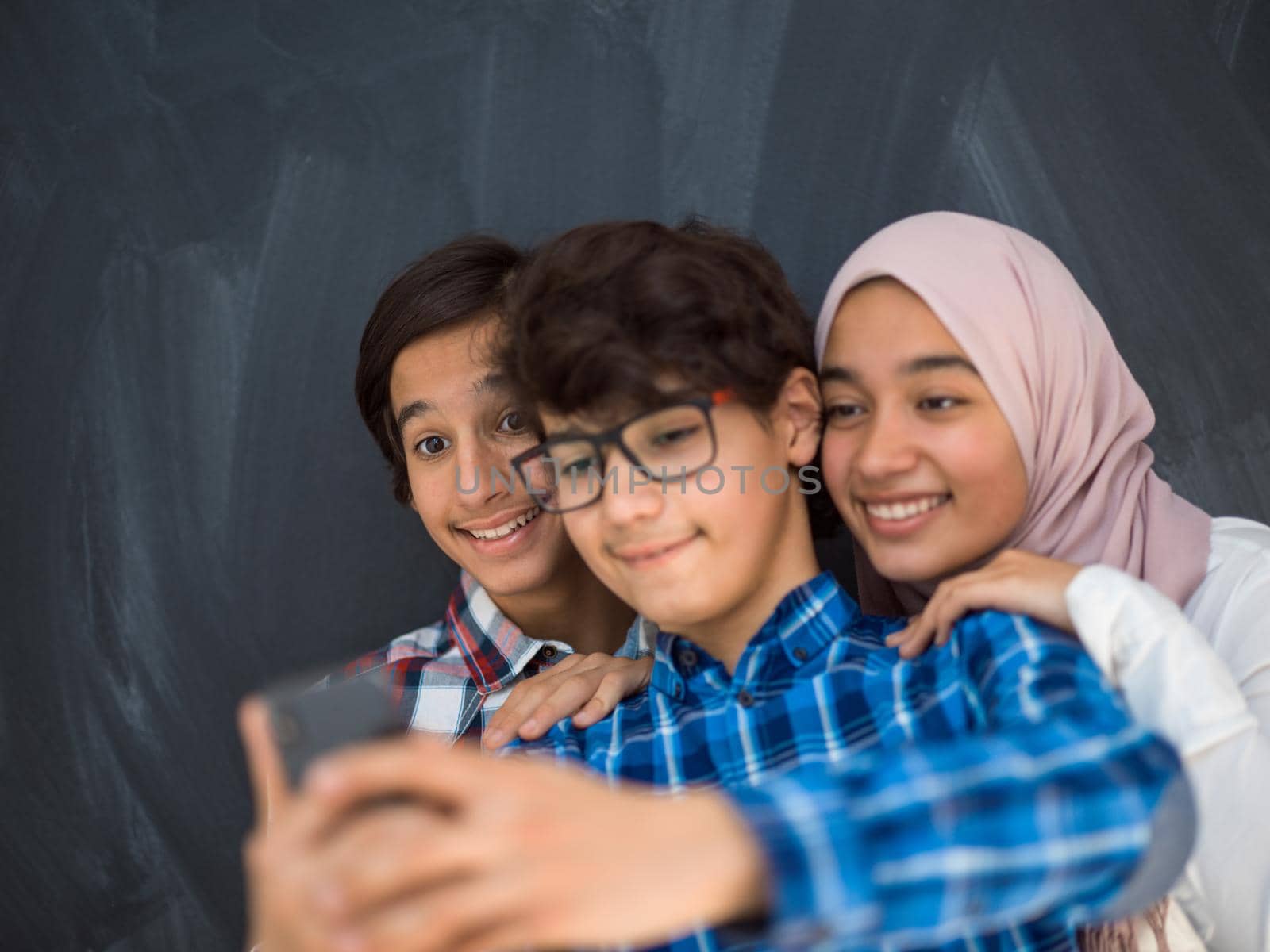 group of arab teens taking selfie photo on smart phone with black chalkboard in background by dotshock