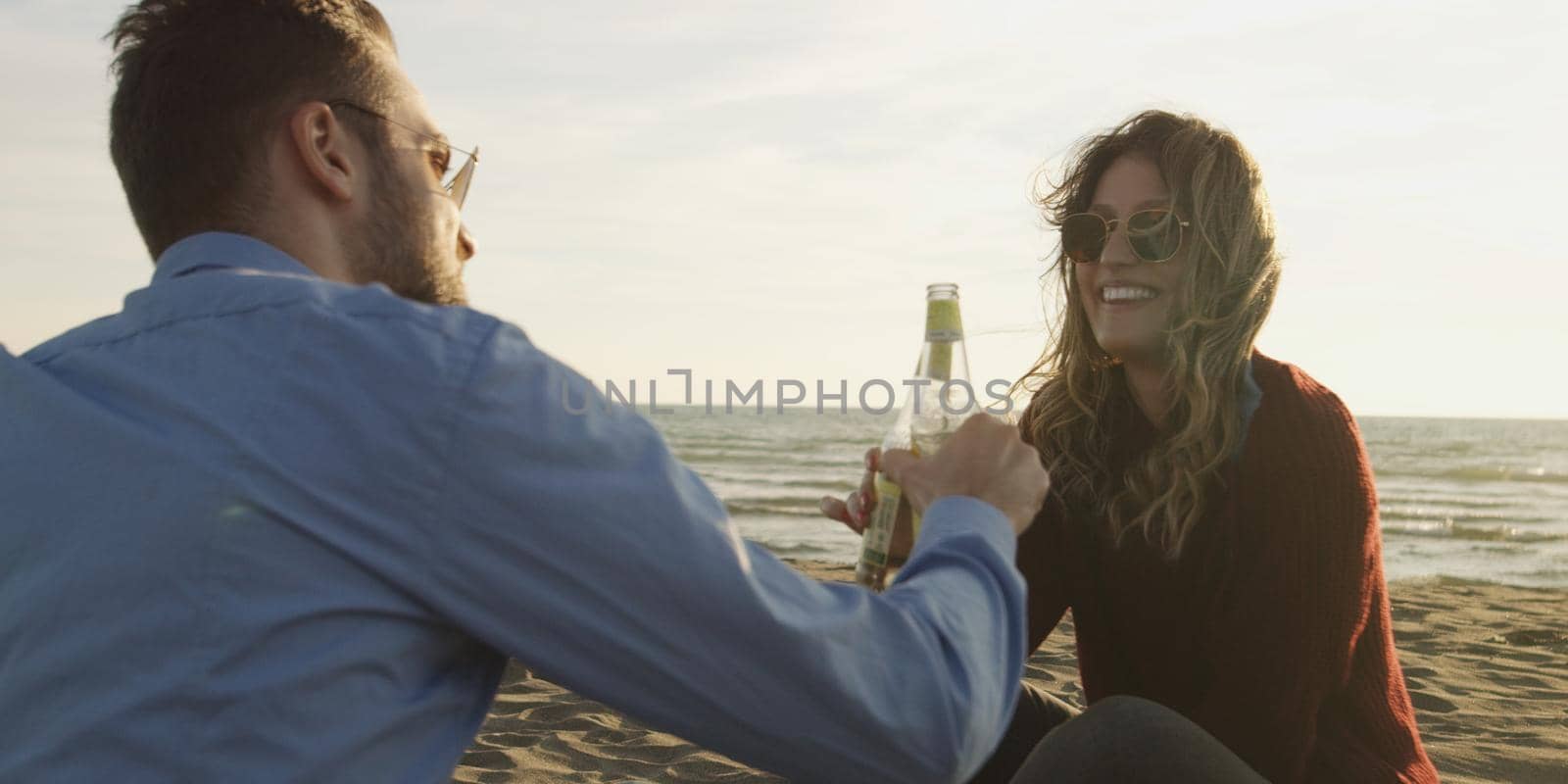 Young Couple Relaxing By The Fire, Drinking A Beer Or A Drink From The Bottle.