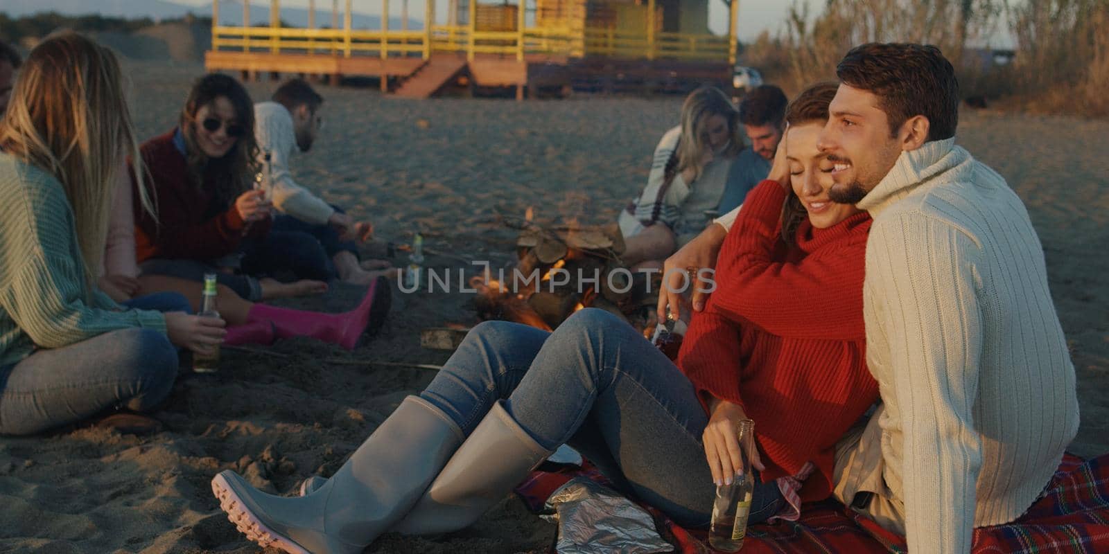 Loving Young Couple Sitting On The Beach beside Campfire drinking beer by dotshock