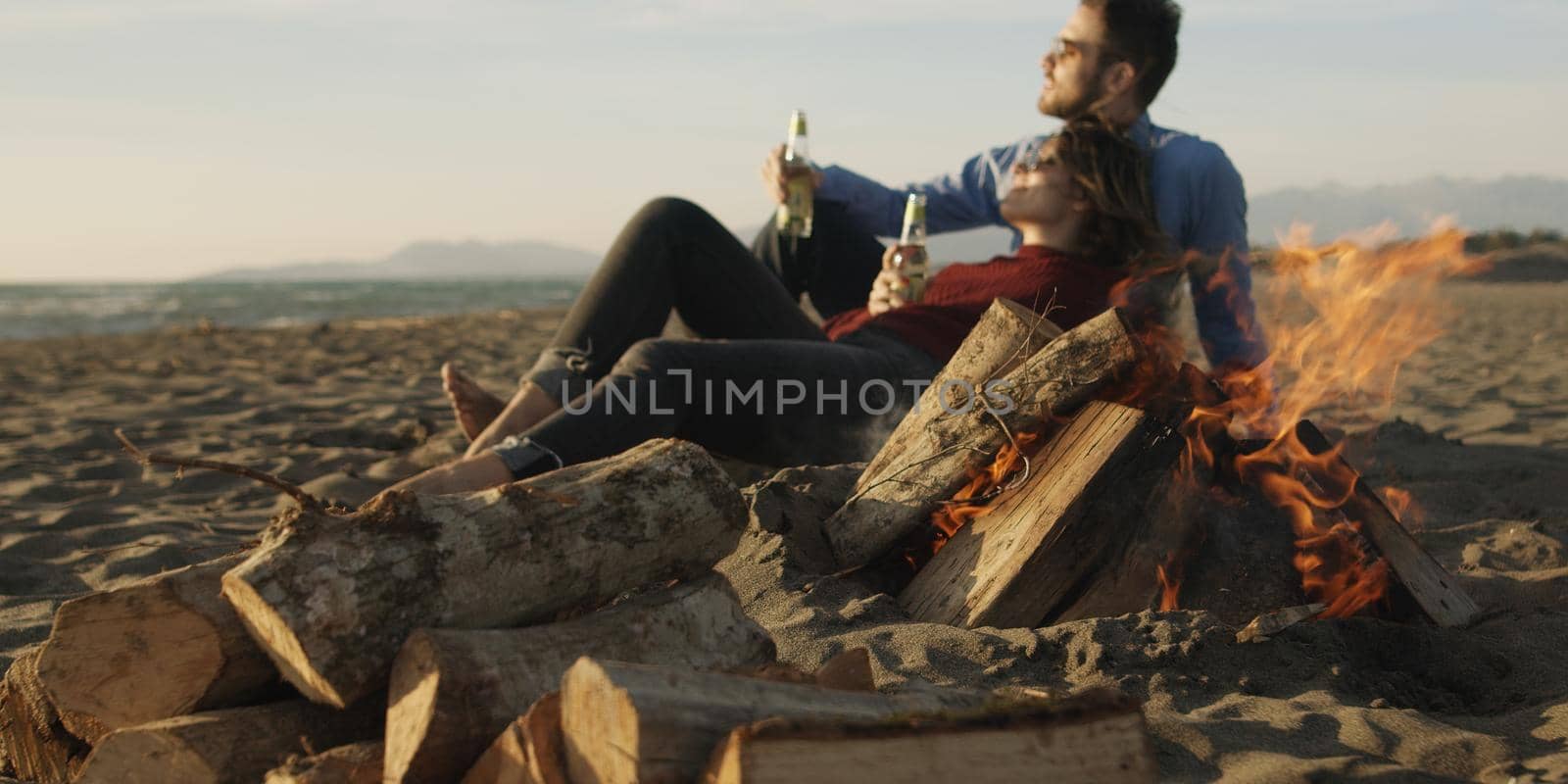 Loving Young Couple Sitting On The Beach beside Campfire drinking beer by dotshock