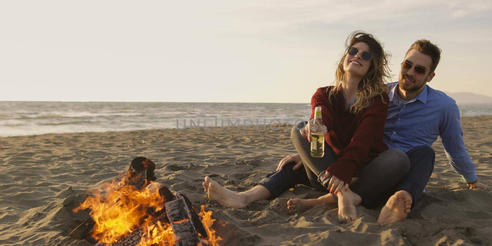 Young Couple Relaxing By The Fire, Drinking A Beer Or A Drink From The Bottle.