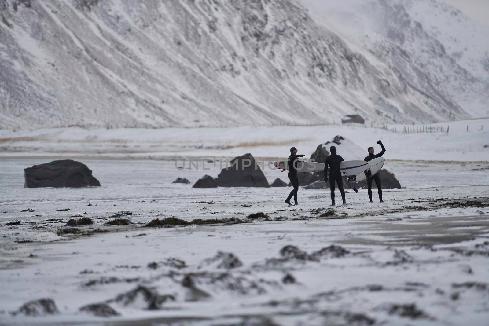 Authentic local Arctic surfers going by beach after surfing in Norwegian sea at snow capped mountain background in winter. Unstad, Norwegian village on Lofoten islands. Norwegian sea coastline. Winter water activities extreme sport