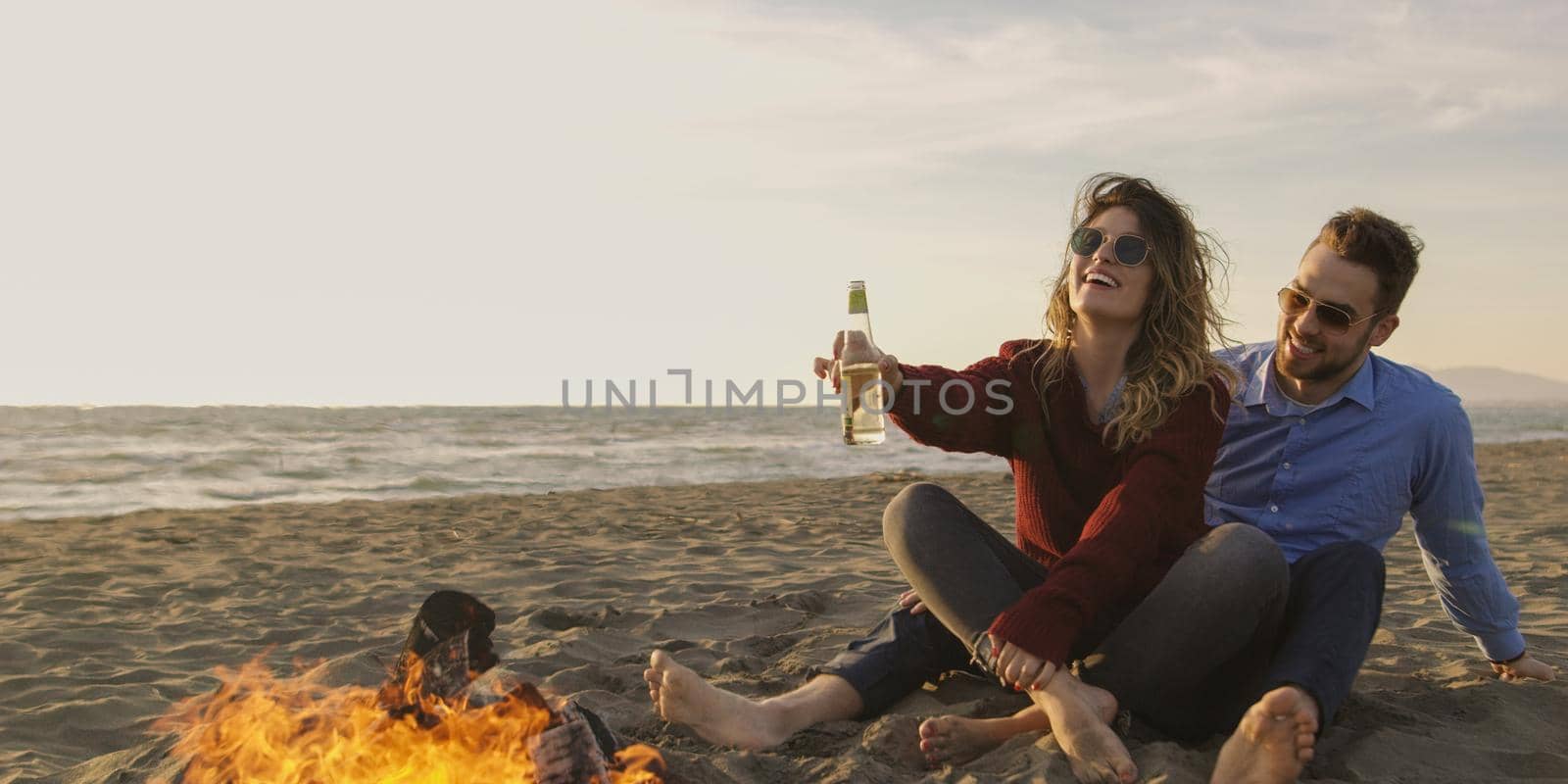 Loving Young Couple Sitting On The Beach beside Campfire drinking beer by dotshock