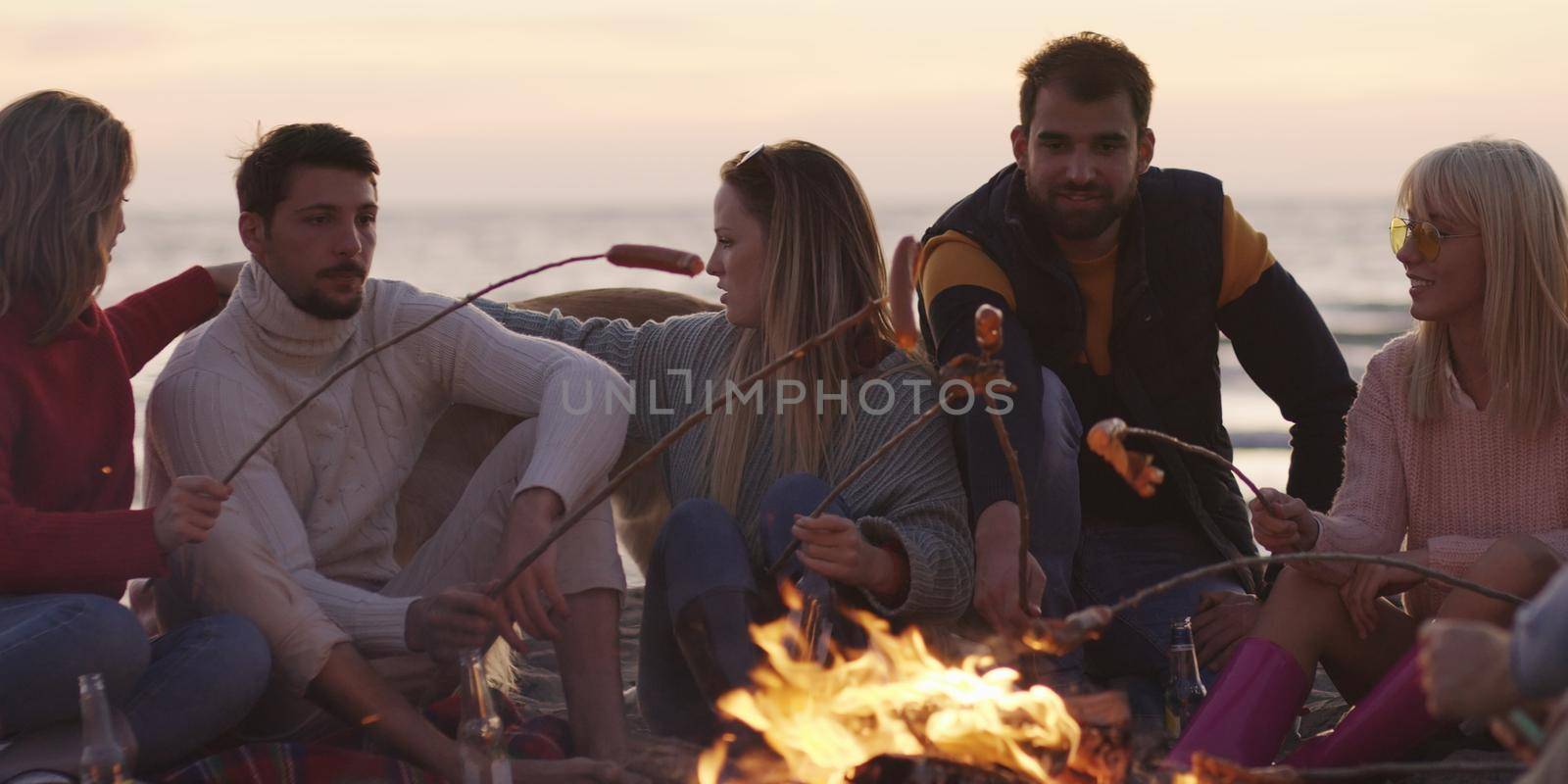 Group of young friends sitting by the fire late at night, grilling sausages and drinking beer, talking and having fun
