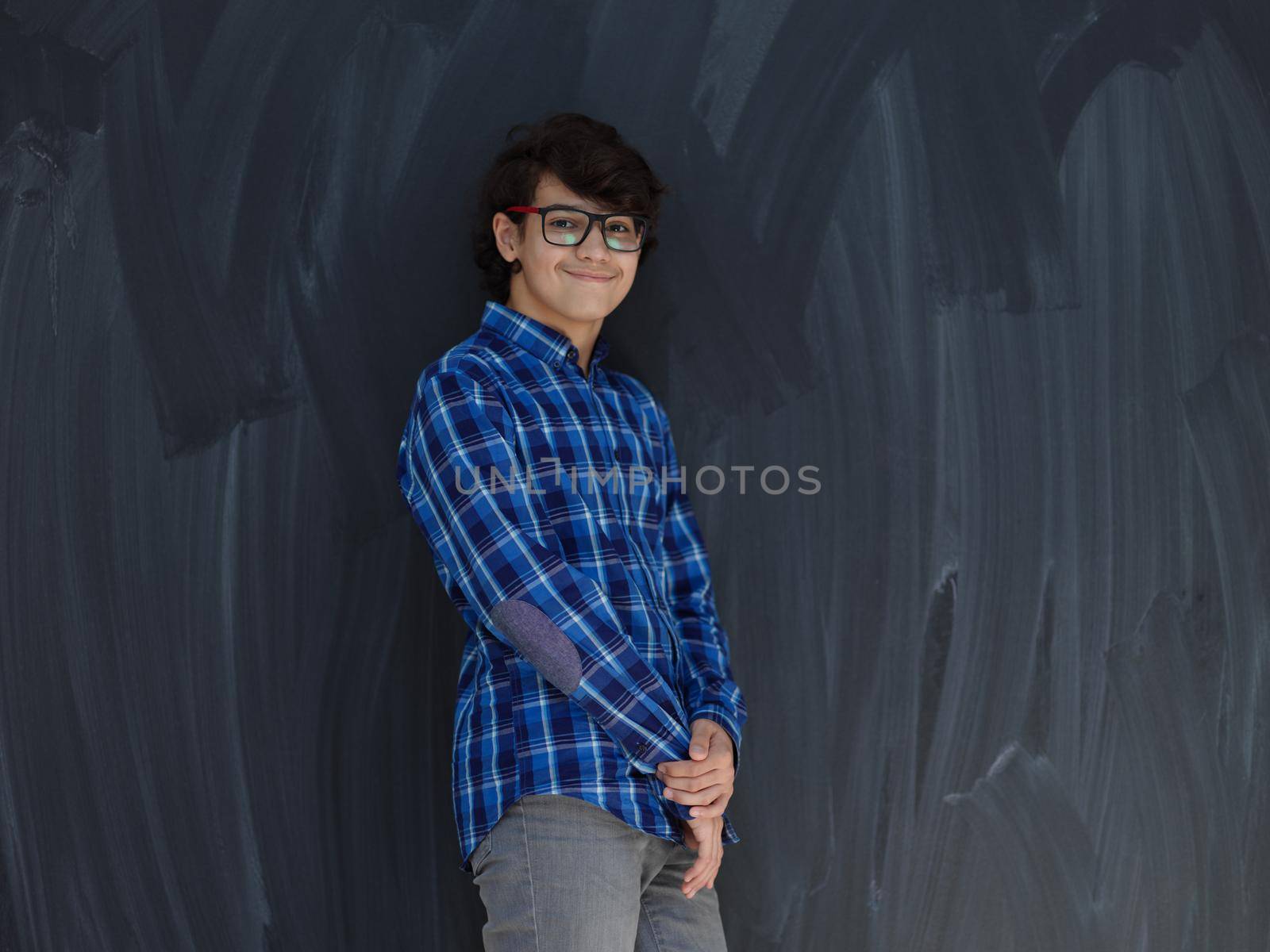 portrait  of smart looking arab teenager with glasses wearing a hat in casual school look against black chalkboard background