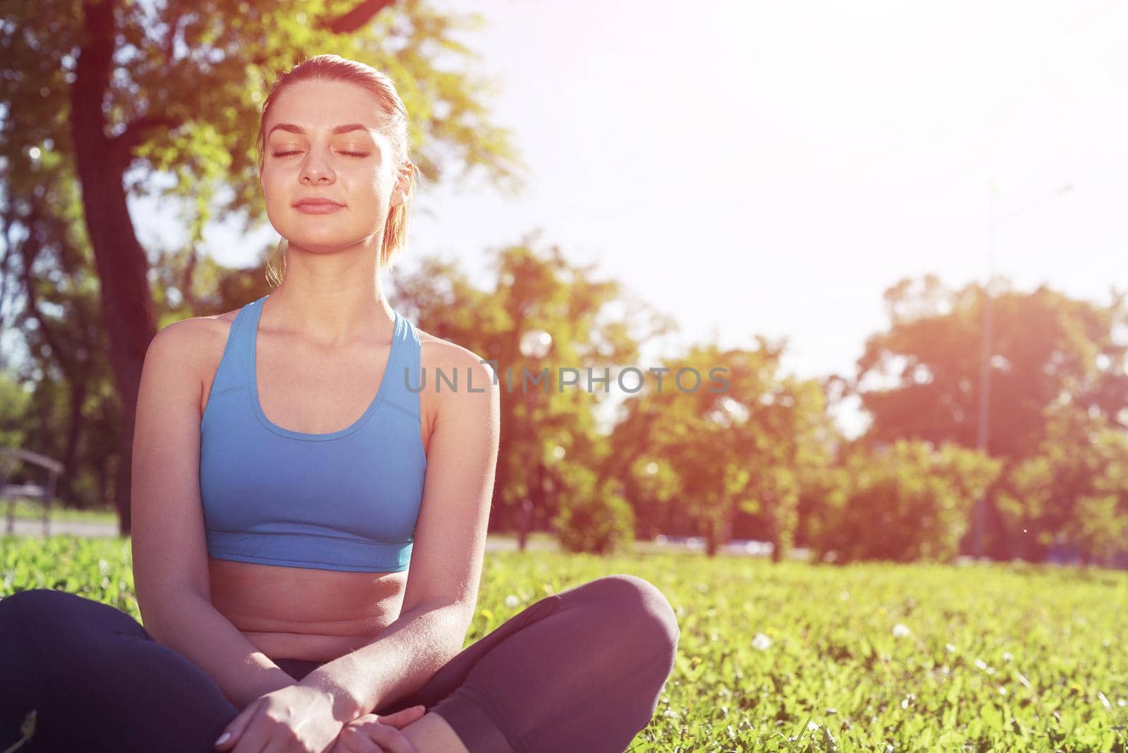 Girl meditates in lotus pose on green grass at sunrise. Practicing of yoga. Portrait of young woman with closed eyes. Training and meditation outdoor at summer day. Healthy lifestyle and relaxation.