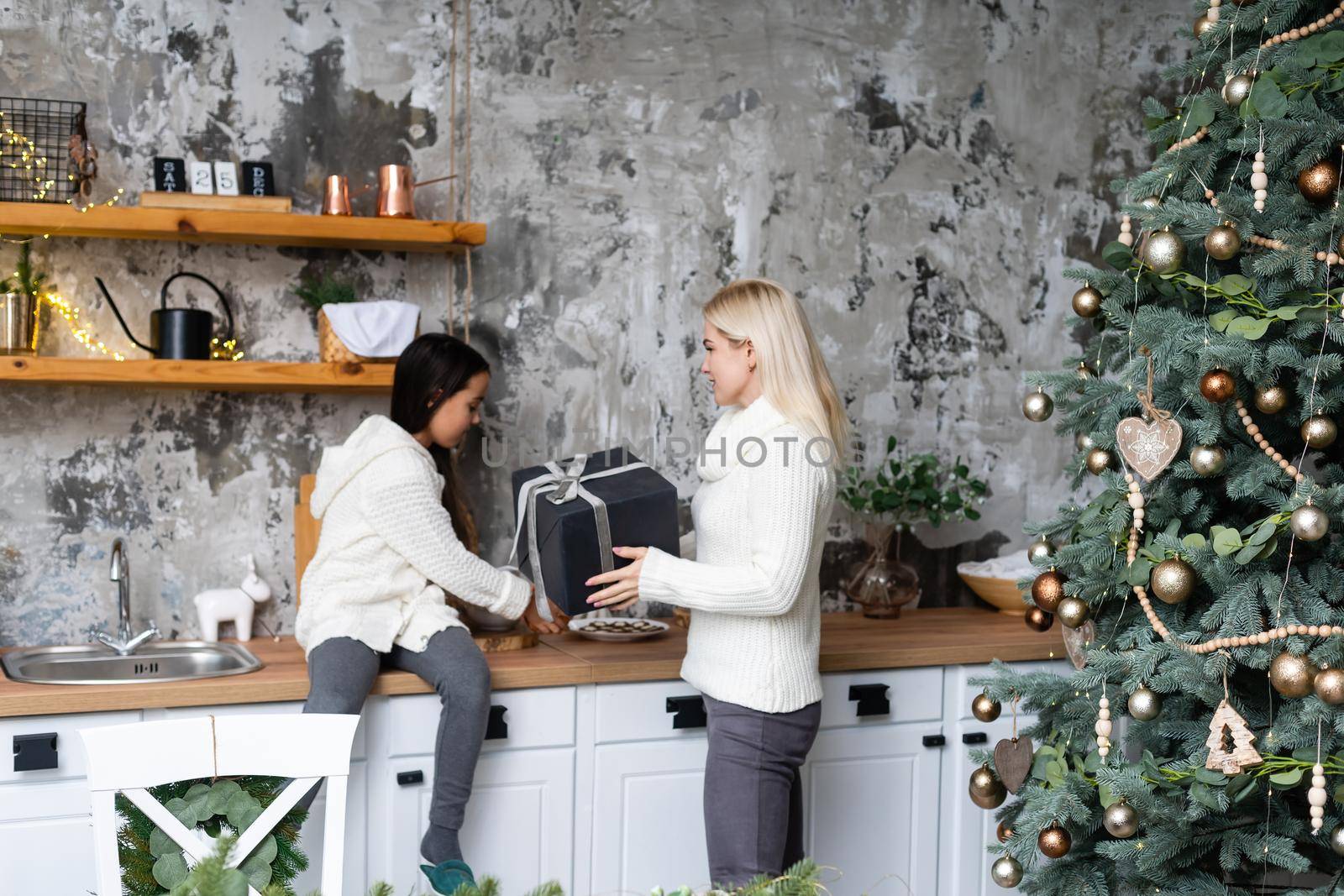 Beautiful mother and daughter have fun, hug and kiss at home near the Christmas tree in a white interior. Family happiness, holiday, joy, vacation, games with a woman. New Year's preparations.