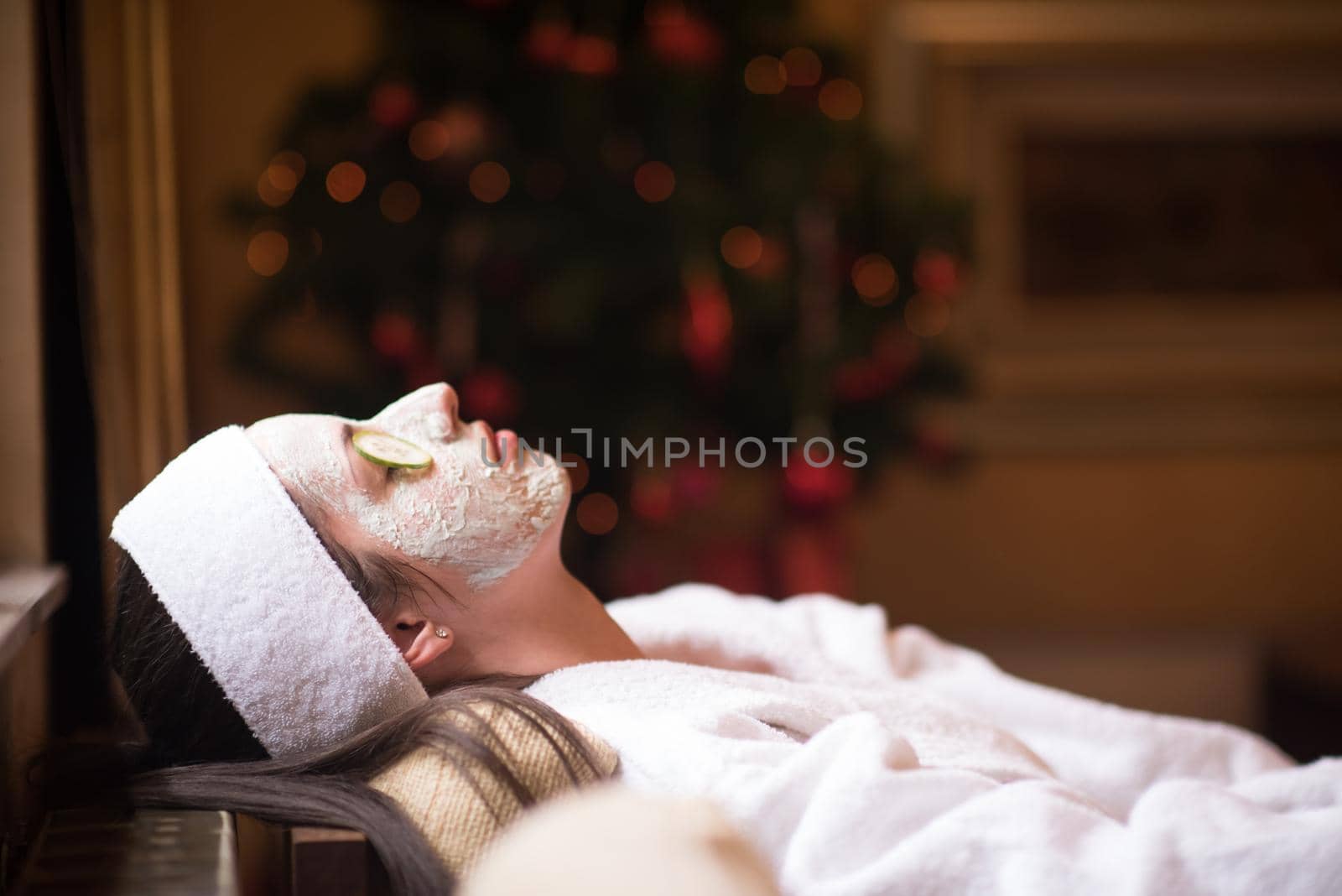 Beautiful young woman is getting facial clay mask at spa, lying with cucumbers on eyes