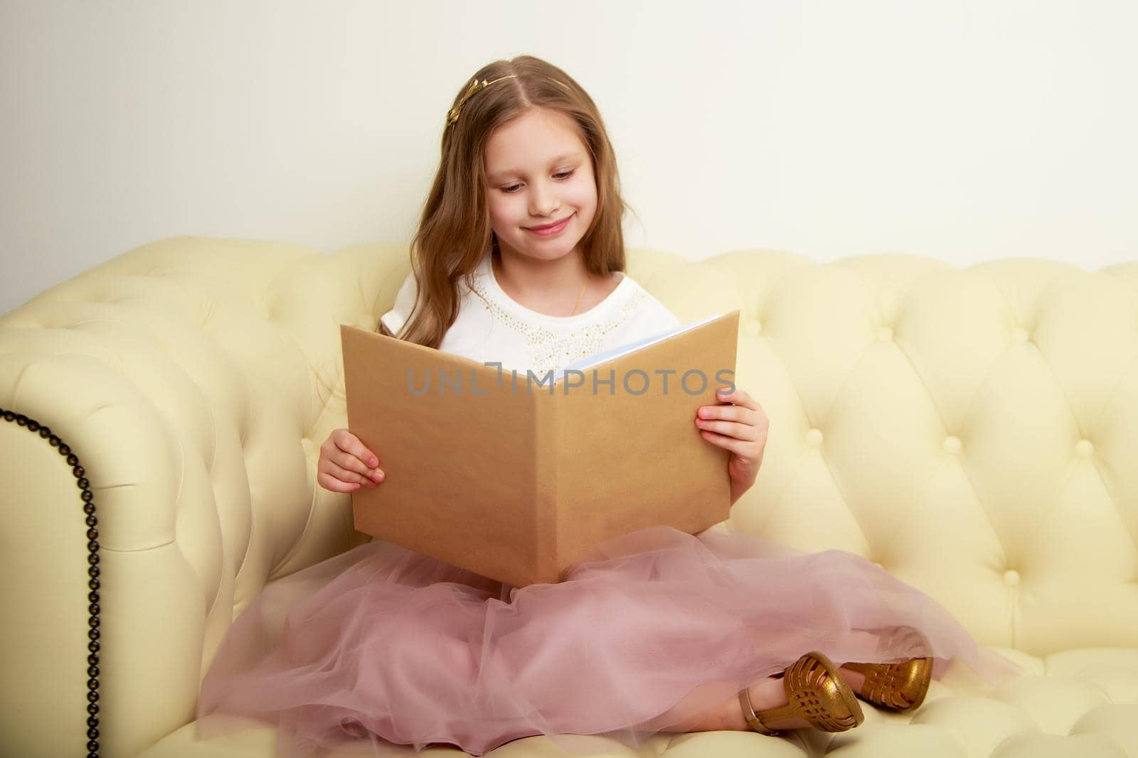 A cute little girl is sitting on the couch and reading a book. The concept of literature, school education and intellectual development of the child.