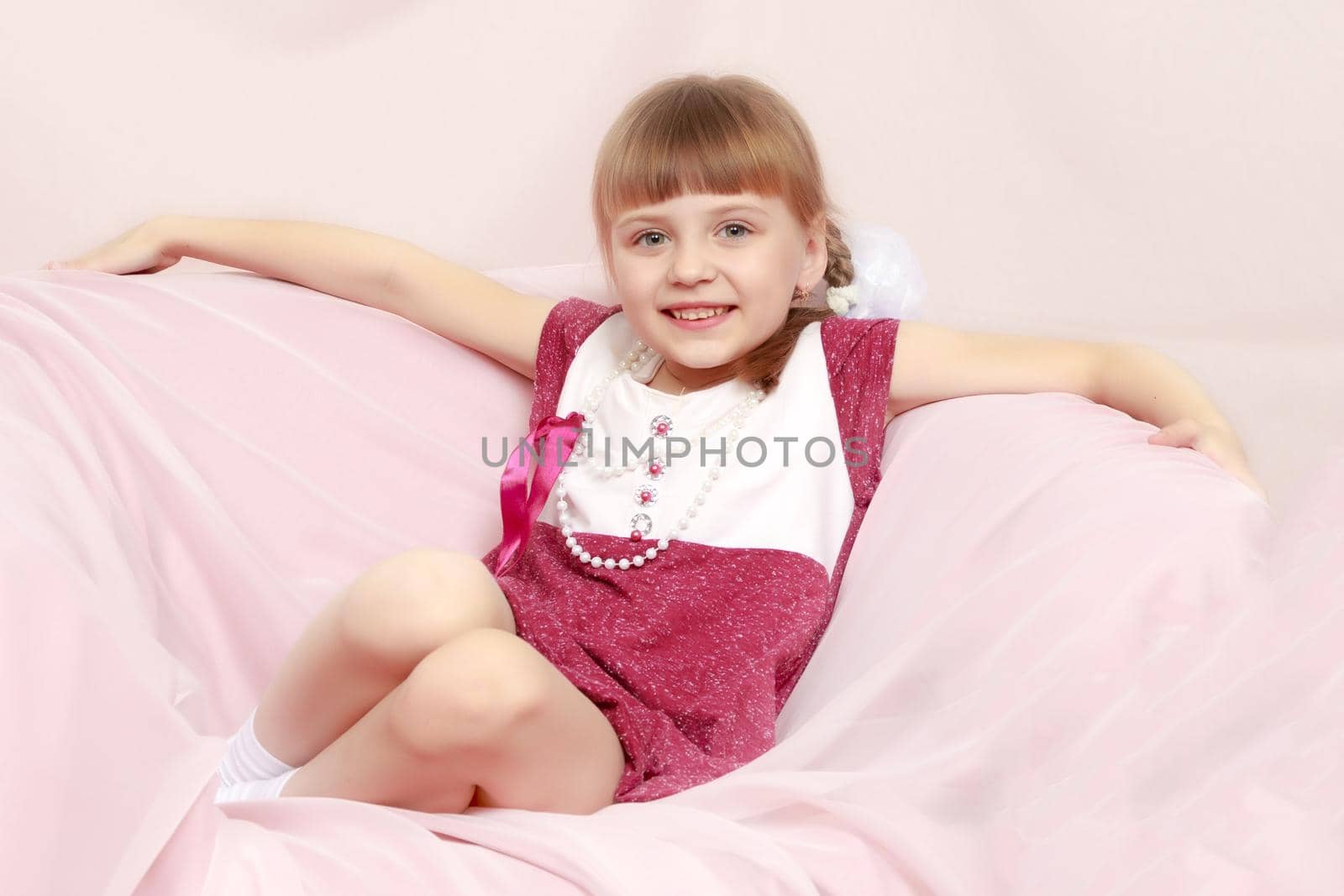 Beautiful little blonde girl with short bangs and pigtails on her head in a good mood.She sits on a pink sofa.