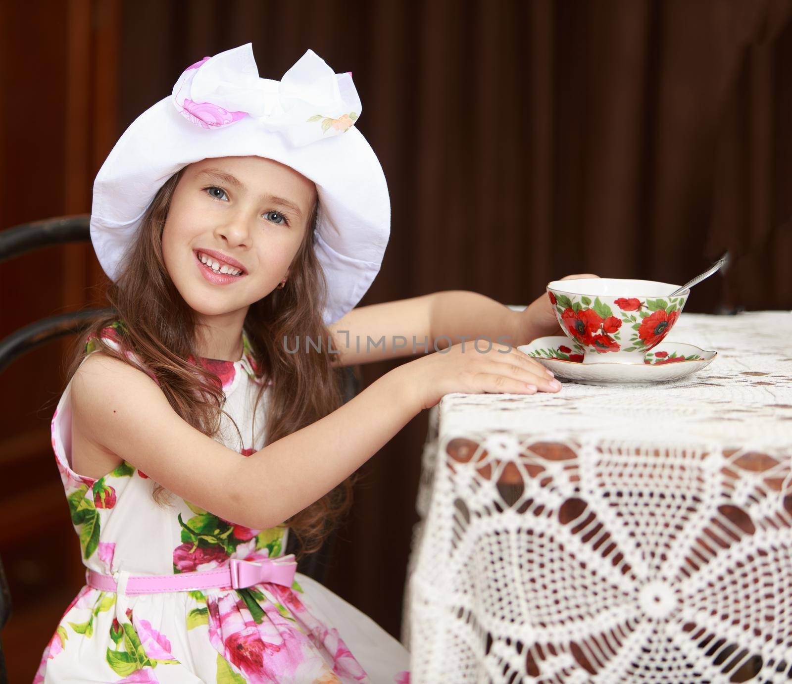 Gentle little girl in a white dress with roses and white hat drinking tea. Girl sitting on an old Viennese chair behind the old oak table , covered with a lace tablecloth. Retro style . close-up