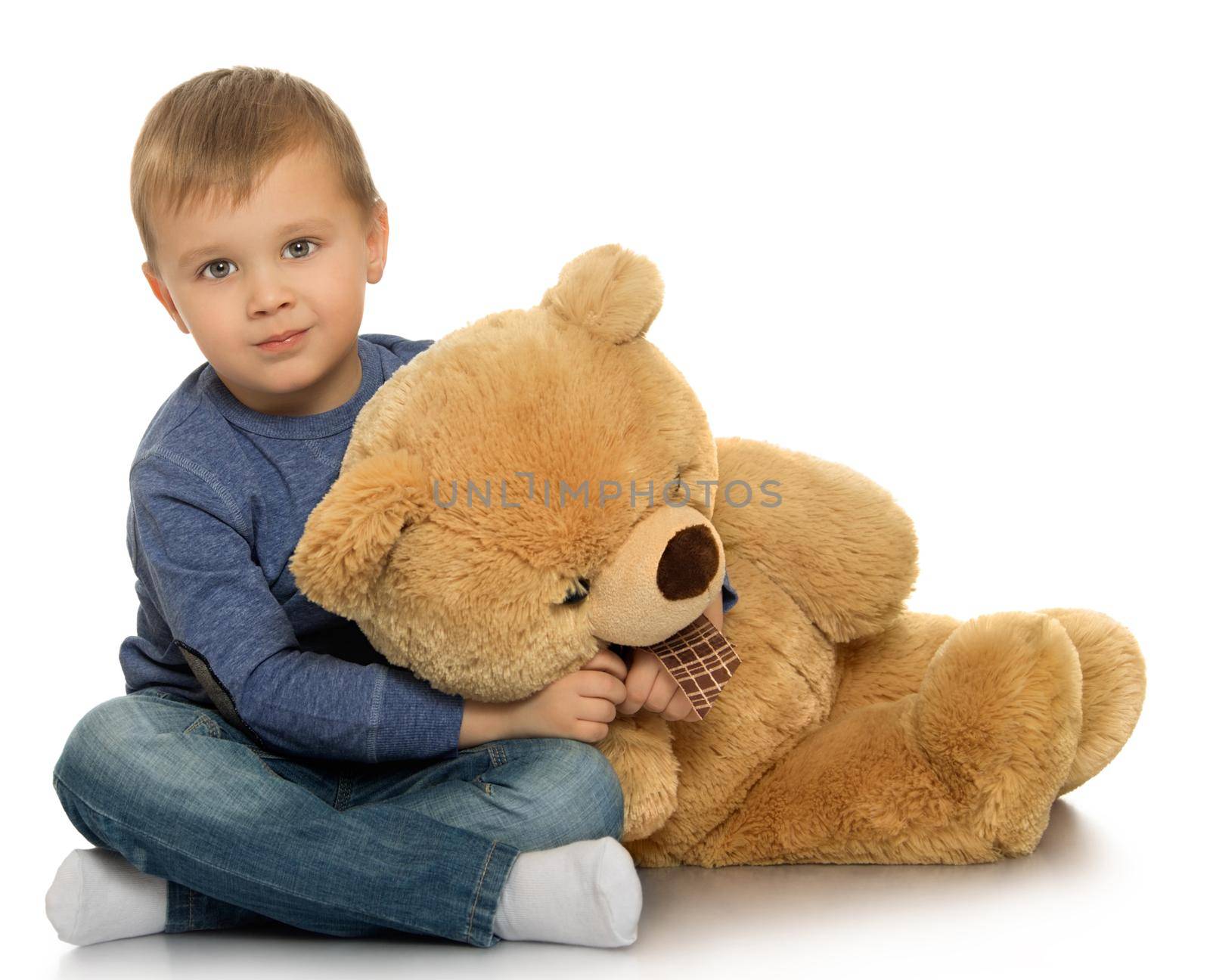 Beautiful little boy sitting on the floor. The boy holds by the neck a large Teddy bear - Isolated on white background