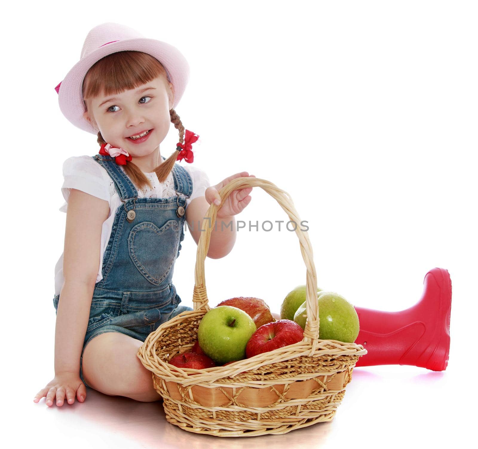 Joyful little girl with a basket which contains apples . Girl sitting on the floor . on the head of the girls pink hat-Isolated on white background