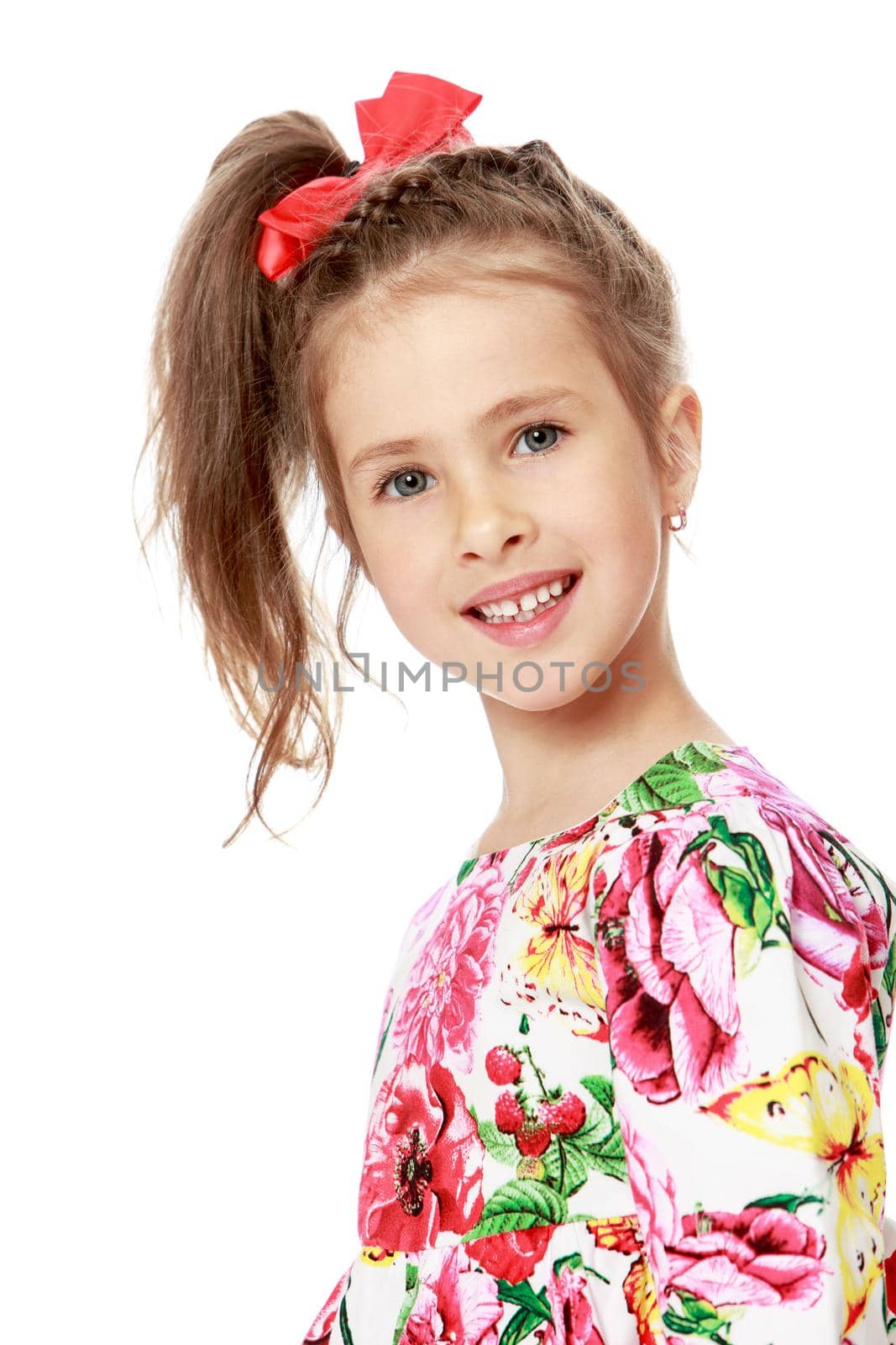 Gentle little girl with a long ponytail down to his shoulders, and a red bow on her head, in a beautiful summer dress. Girl smiling. Close-up - Isolated on white background