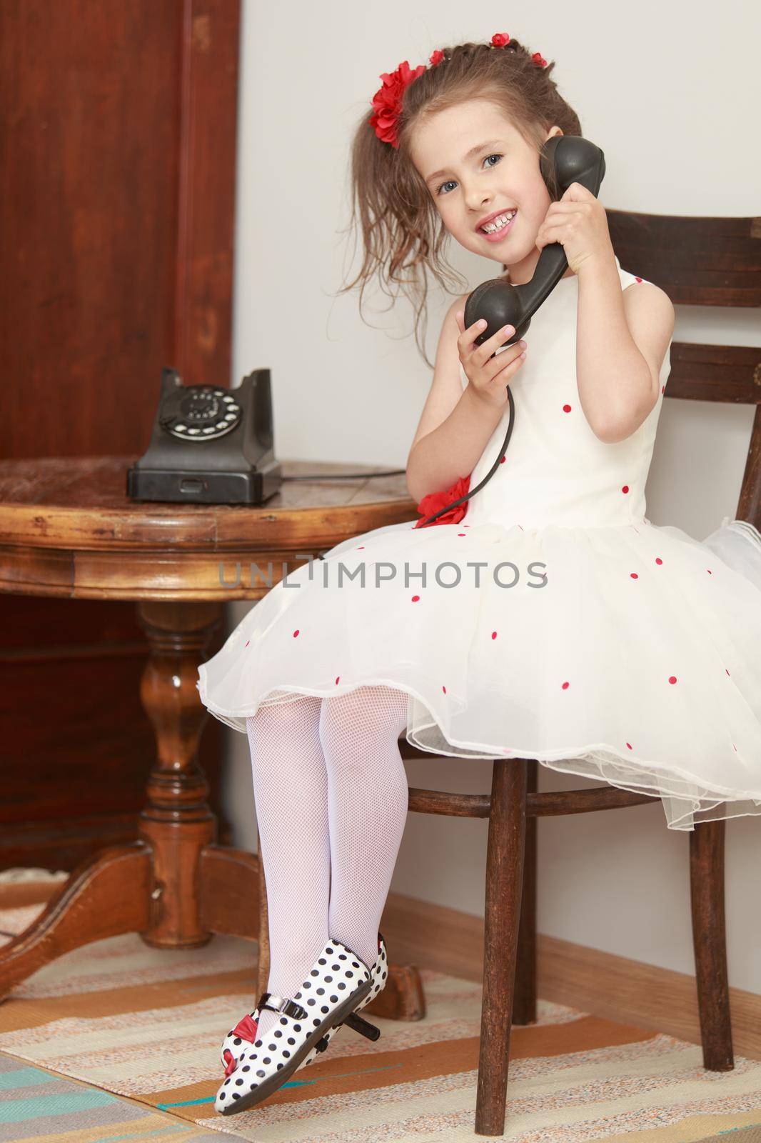 Shy little girl sitting on an old Viennese chair beside a round oak table. Girl holding the ear up the old phone
