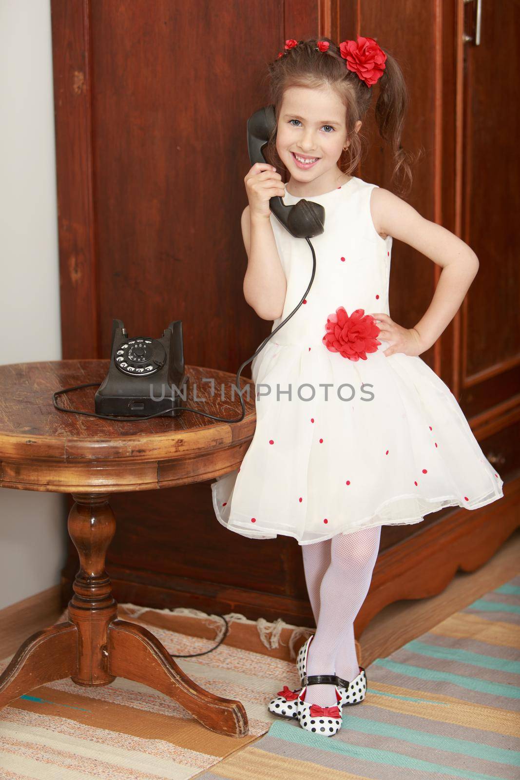 Cute little girl stands beside the round oak table. Girl holding the ear up the old phone