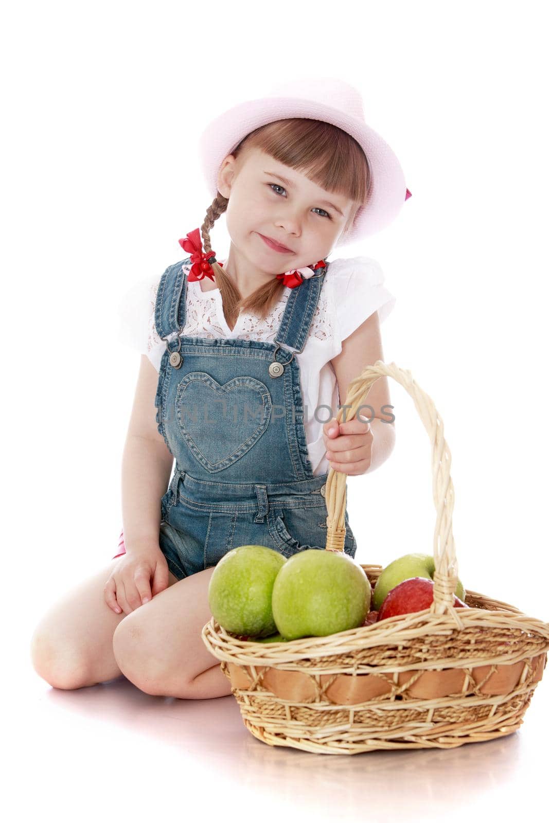 Joyful little girl with a basket which contains apples . Girl sitting on the floor . on the head of the girls pink hat-Isolated on white background
