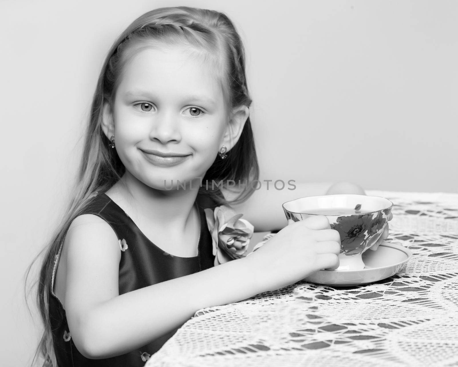 A beautiful little girl with long blond hair sitting at a table and drinking tea.Black and white photography.