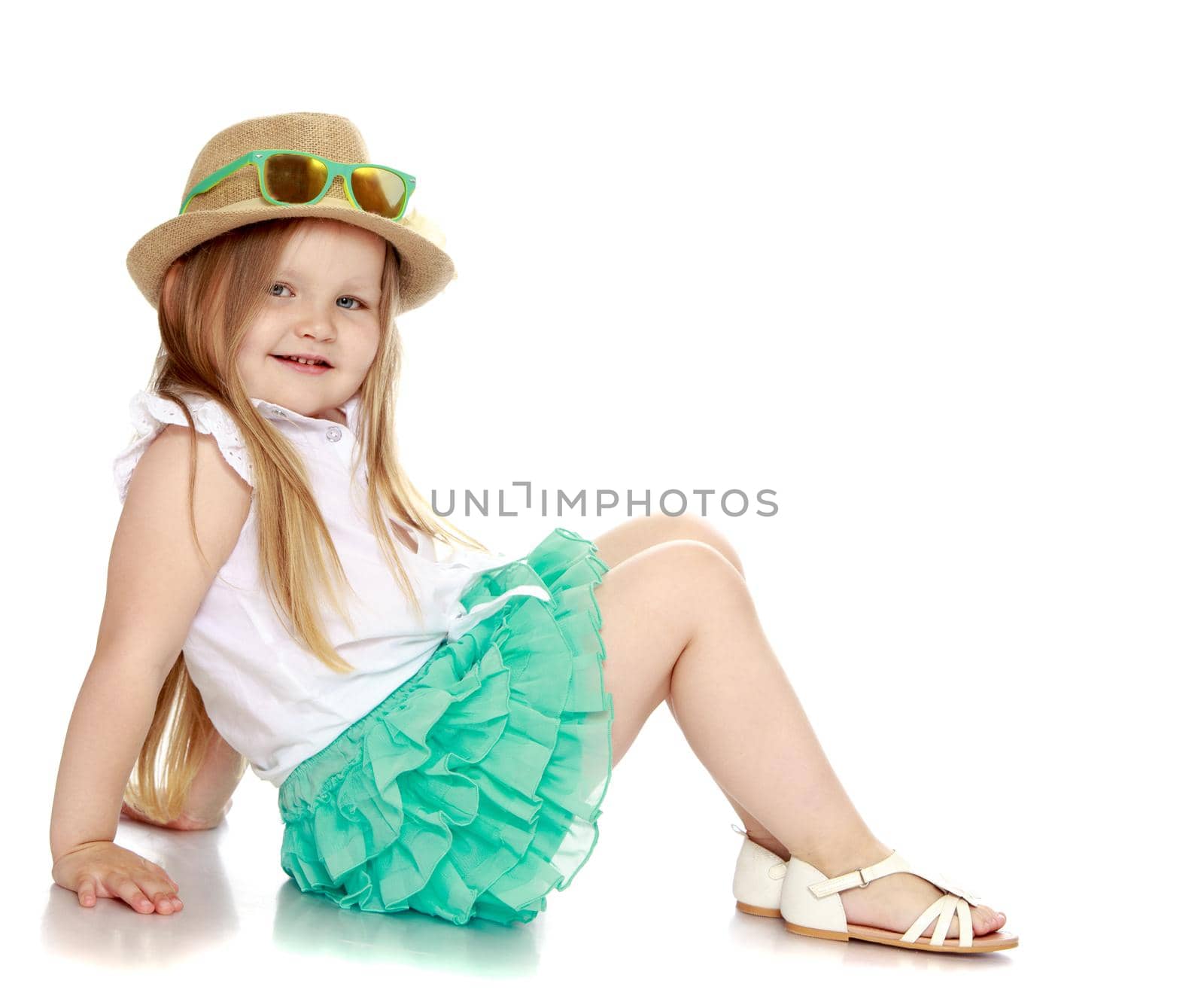 Caucasian little girl with long blond hair below the shoulders , a hat and sunglasses and a short green skirt . Girl sitting on the floor turned sideways to the camera - Isolated on white background