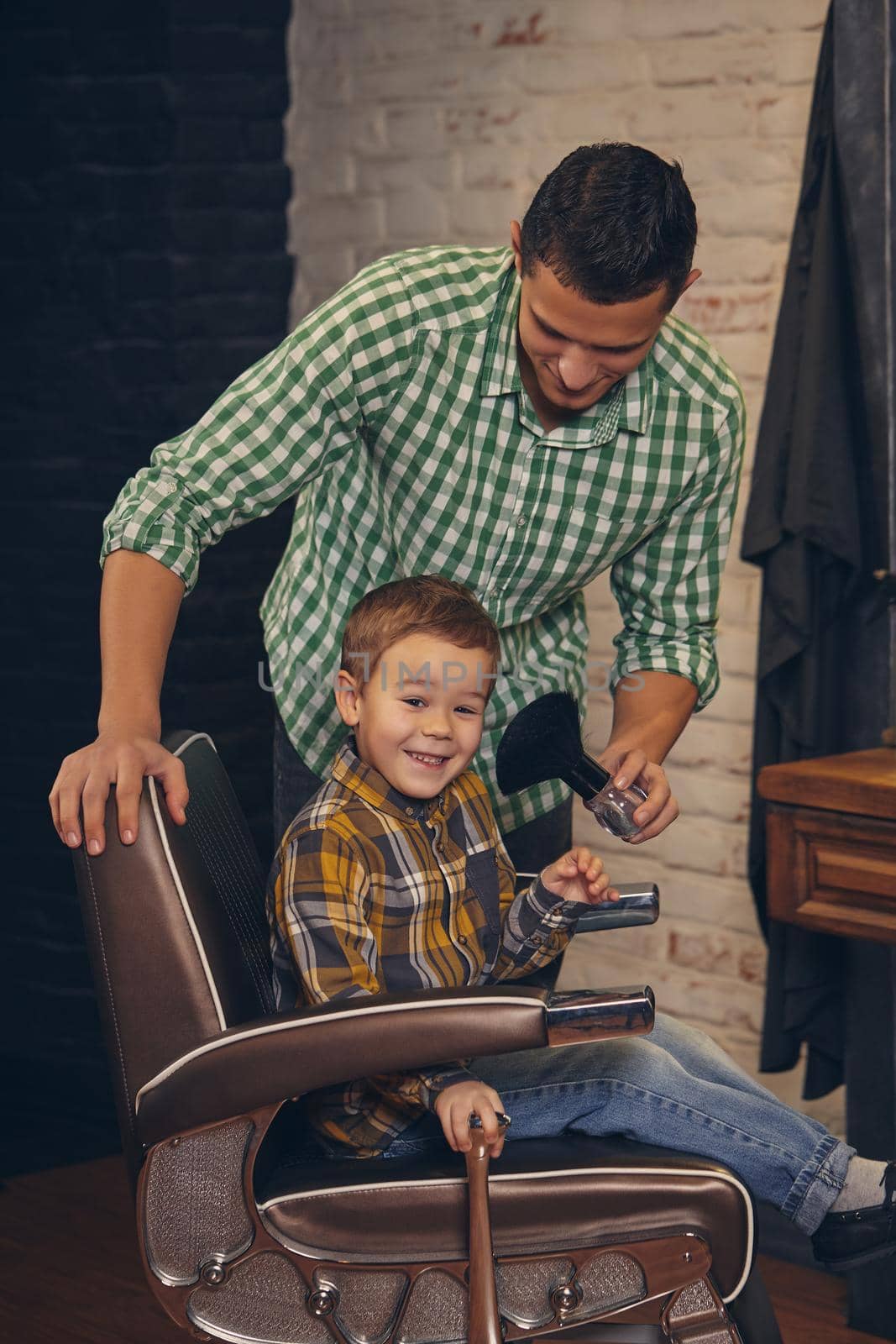stylish little kid sitting on chair at barbershop with his young father on background, they are fooling around and having fun