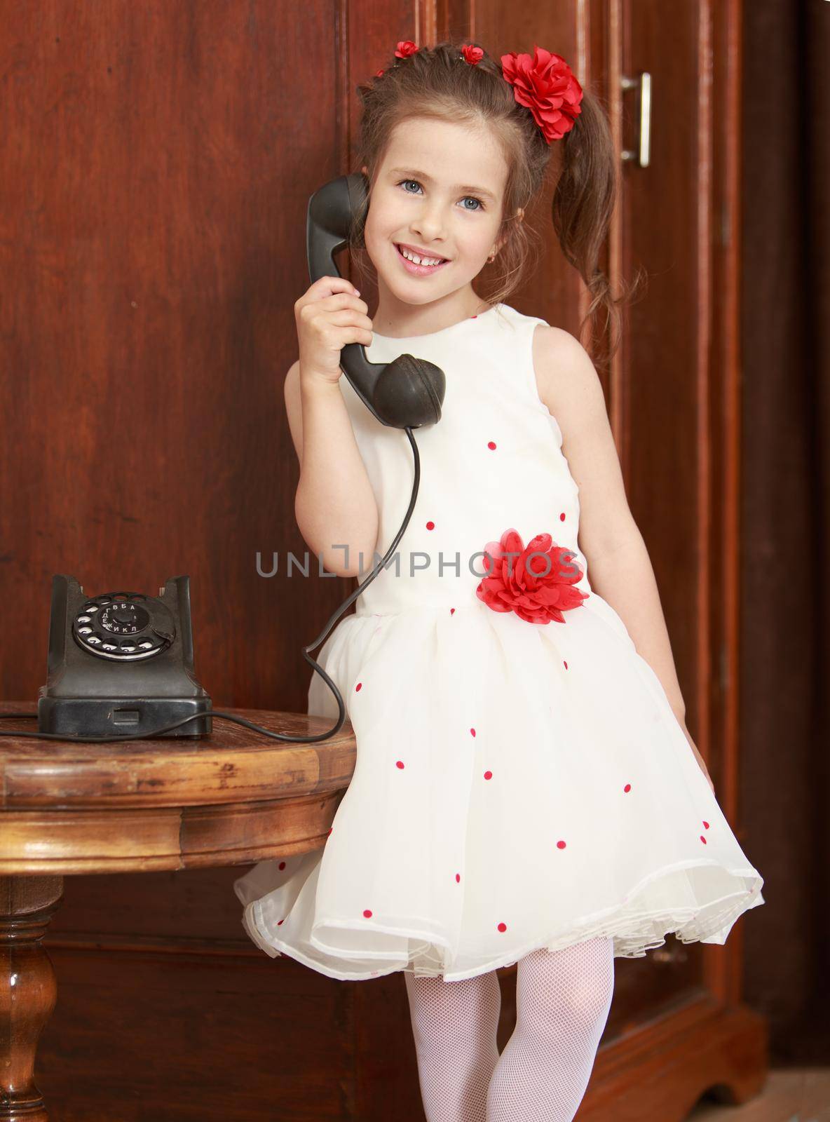 Cute little girl stands beside the round oak table. Girl holding the ear up the old phone