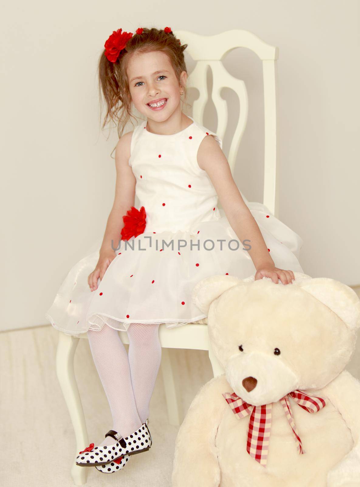 Cute little girl in white dress sitting on a white chair . The girl holds the head of a large Teddy bear