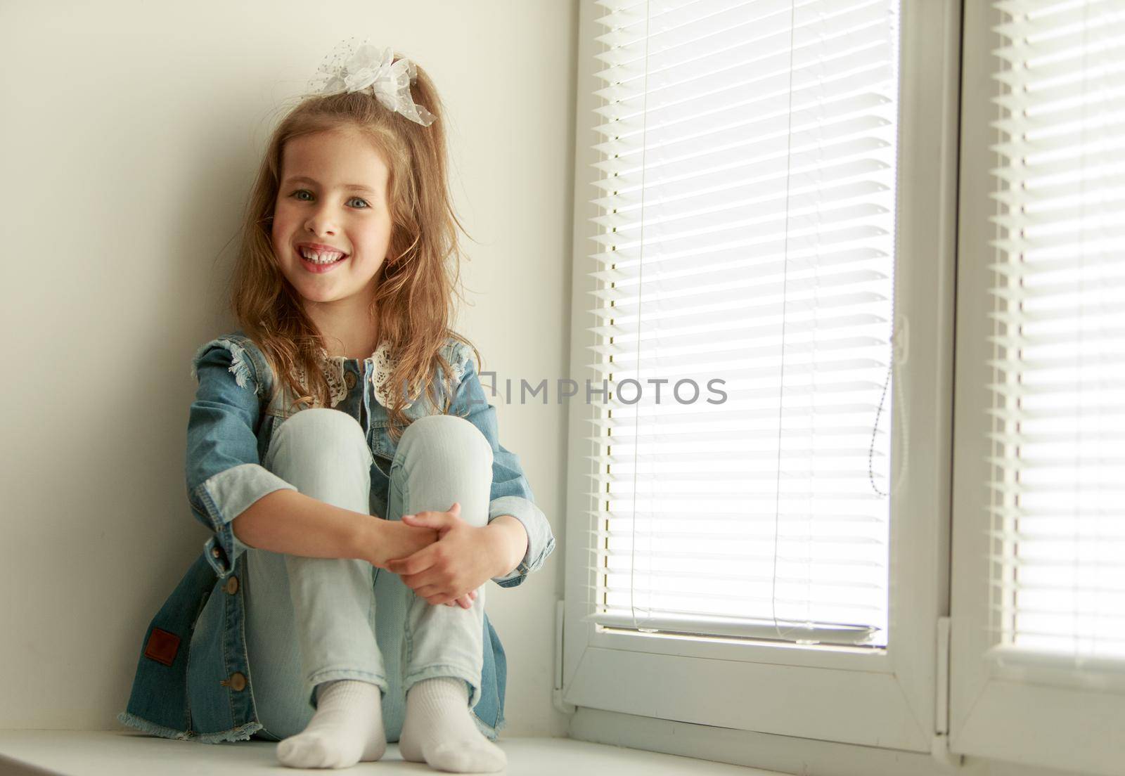 Cute little girl sitting on the windowsill near the window , around the closed blinds. The girl hugged her knees