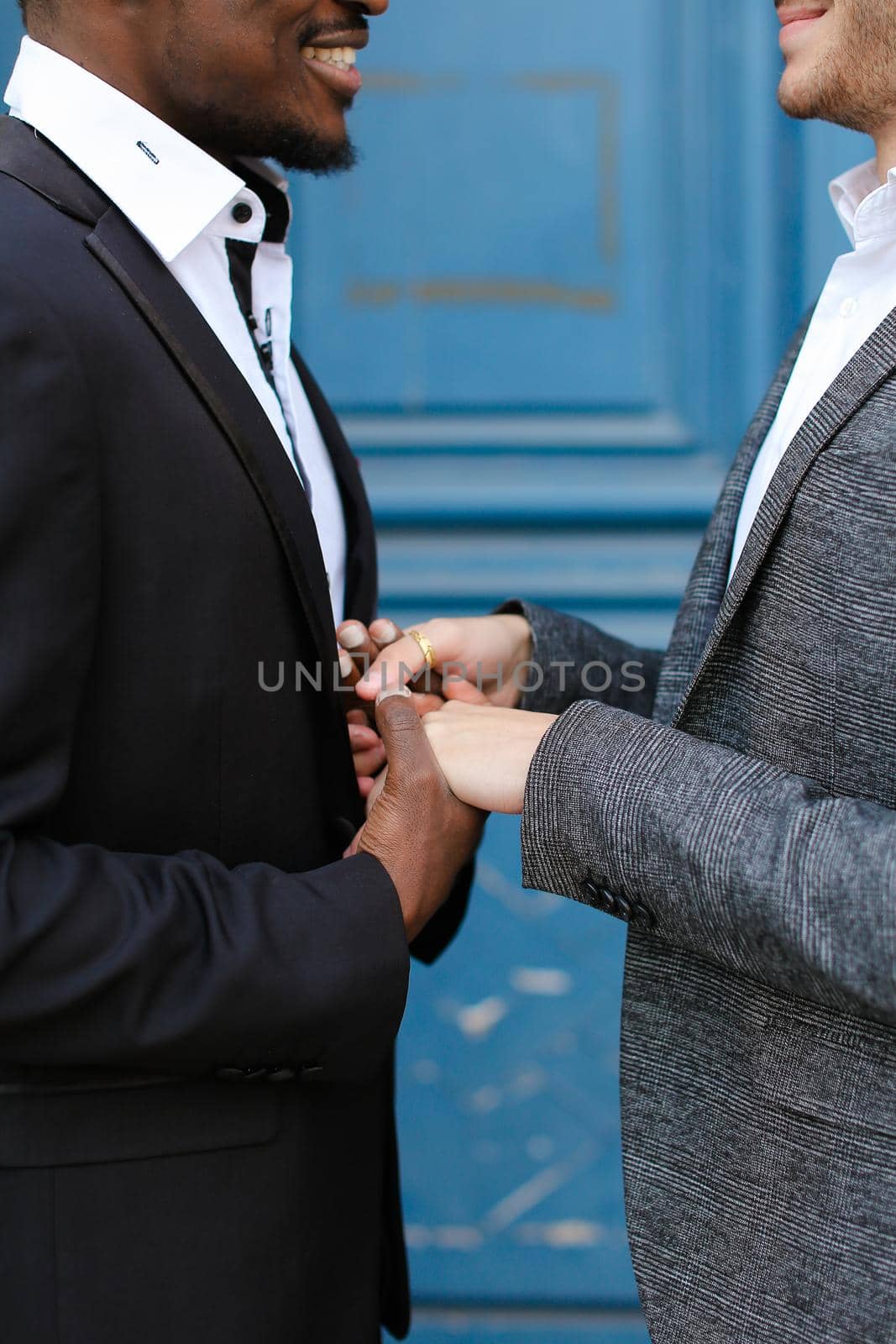 Afro american smiling man holding hands of caucasian guy in door background, wearing suit. Concept of same sex couple and gays.