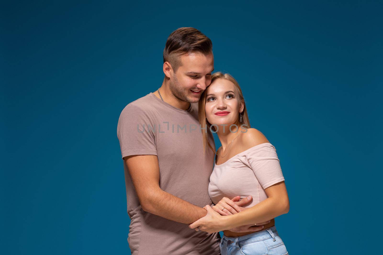 Young couple is hugging on blue background in studio. They wear T-shirts, jeans and smile. by nazarovsergey