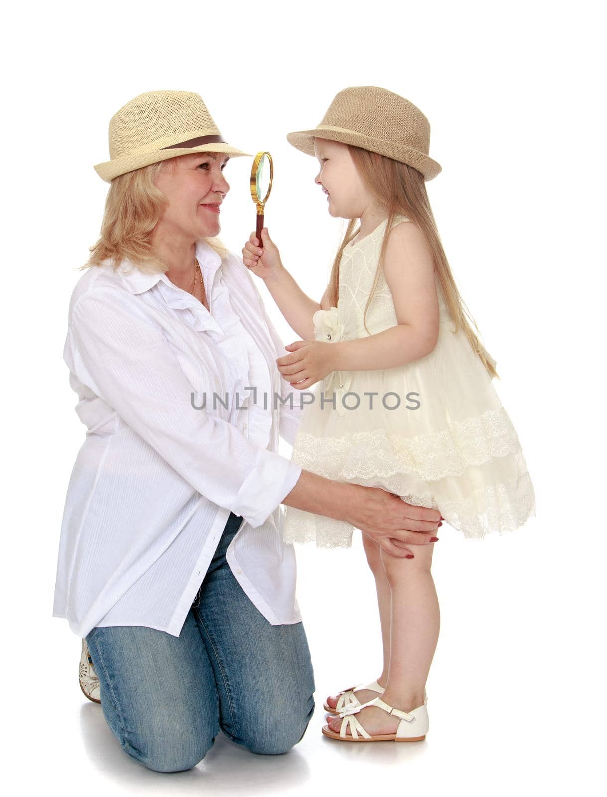 Cute little girl with long blond hair in white dress and hat . The girl looks at through a magnifying glass the face of his beloved grandmother - Isolated on white background