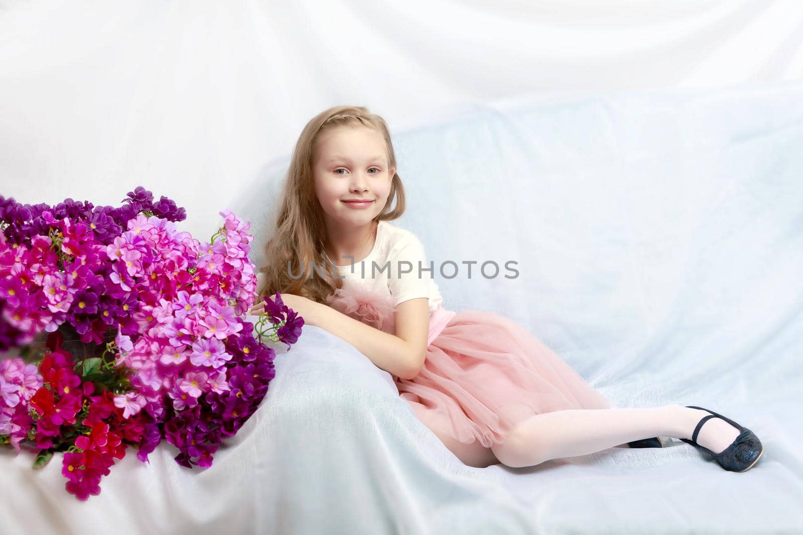 Caucasian little girl with long blond hair, in a beautiful pink skirt and white T-shirt and a pink bow on her chest.Sits on a blue sofa.A bouquet of pink flowers.