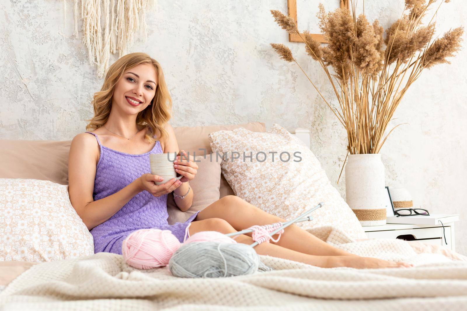 Young woman holding a mug with tea or coffee sitting on the bed in the bedroom. Near lie yarn and knitting needles. Horizontal photo. Freelance creative working and living concept.