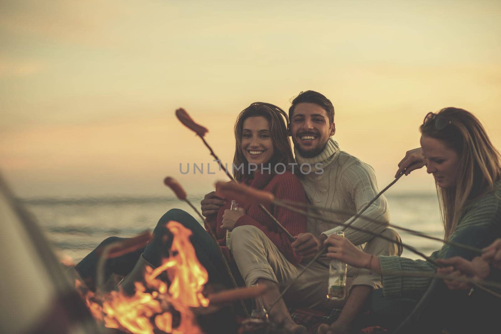 Group of young friends sitting by the fire at autumn beach, grilling sausages and drinking beer, talking and having fun filter